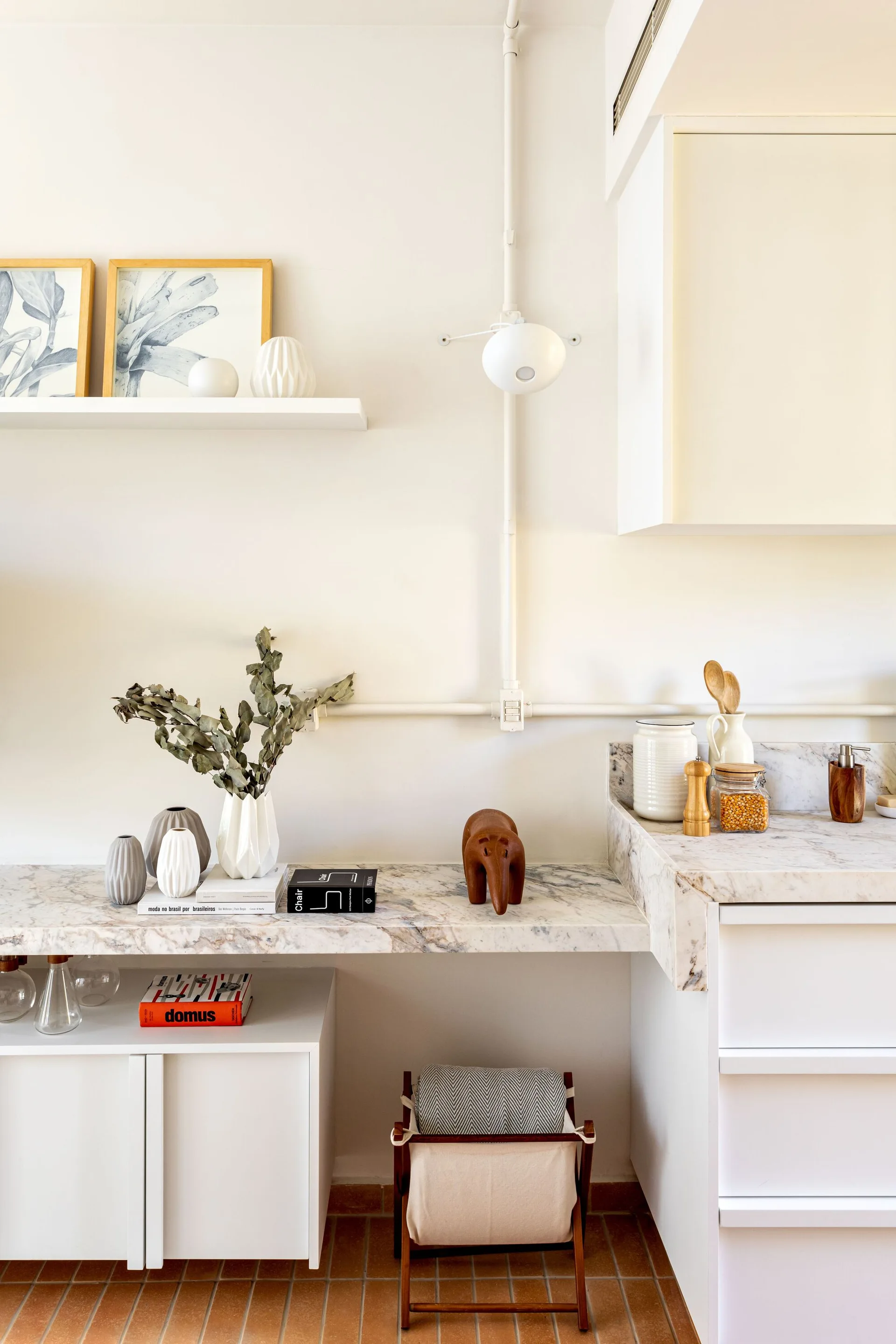 A white minimalist kitchen features a veined marble countertop, modern white cabinetry, terracotta floor tiles, and curated ceramics.