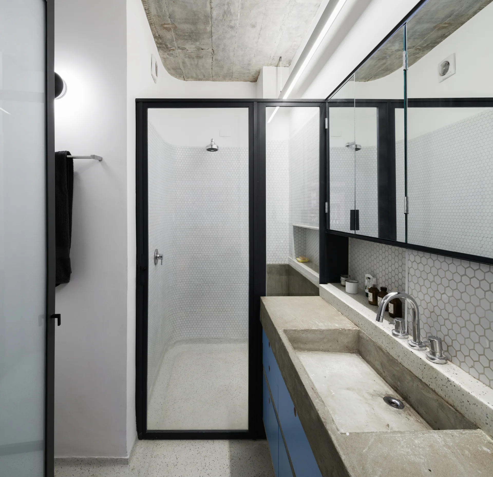 A modern industrial bathroom features a concrete vanity with blue cabinets, a glass shower stall, and white hexagon wall tiles.