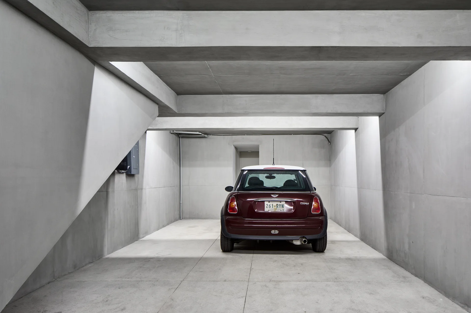 A maroon hatchback car is parked in a minimalist, all-concrete garage featuring thick structural beams and smooth grey walls.