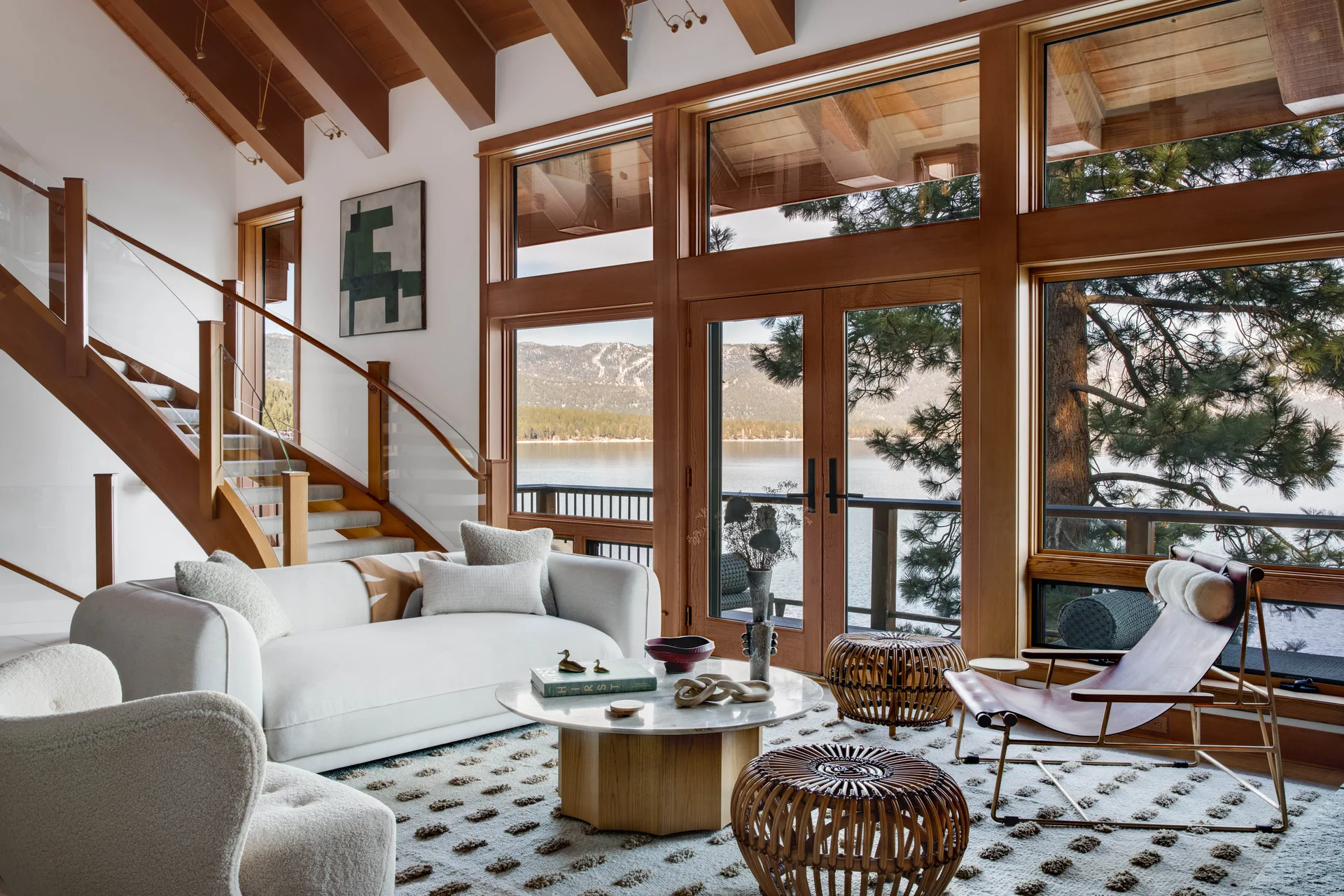 A rustic contemporary living room features a white sofa, a marble coffee table, and large wood-framed windows overlooking a lake.