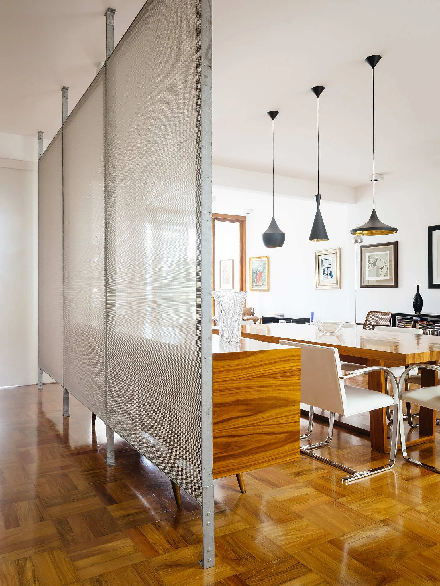 A bright dining area features a floor-to-ceiling metal-framed screen, a polished wood table with white chairs, and three black pendant lights.