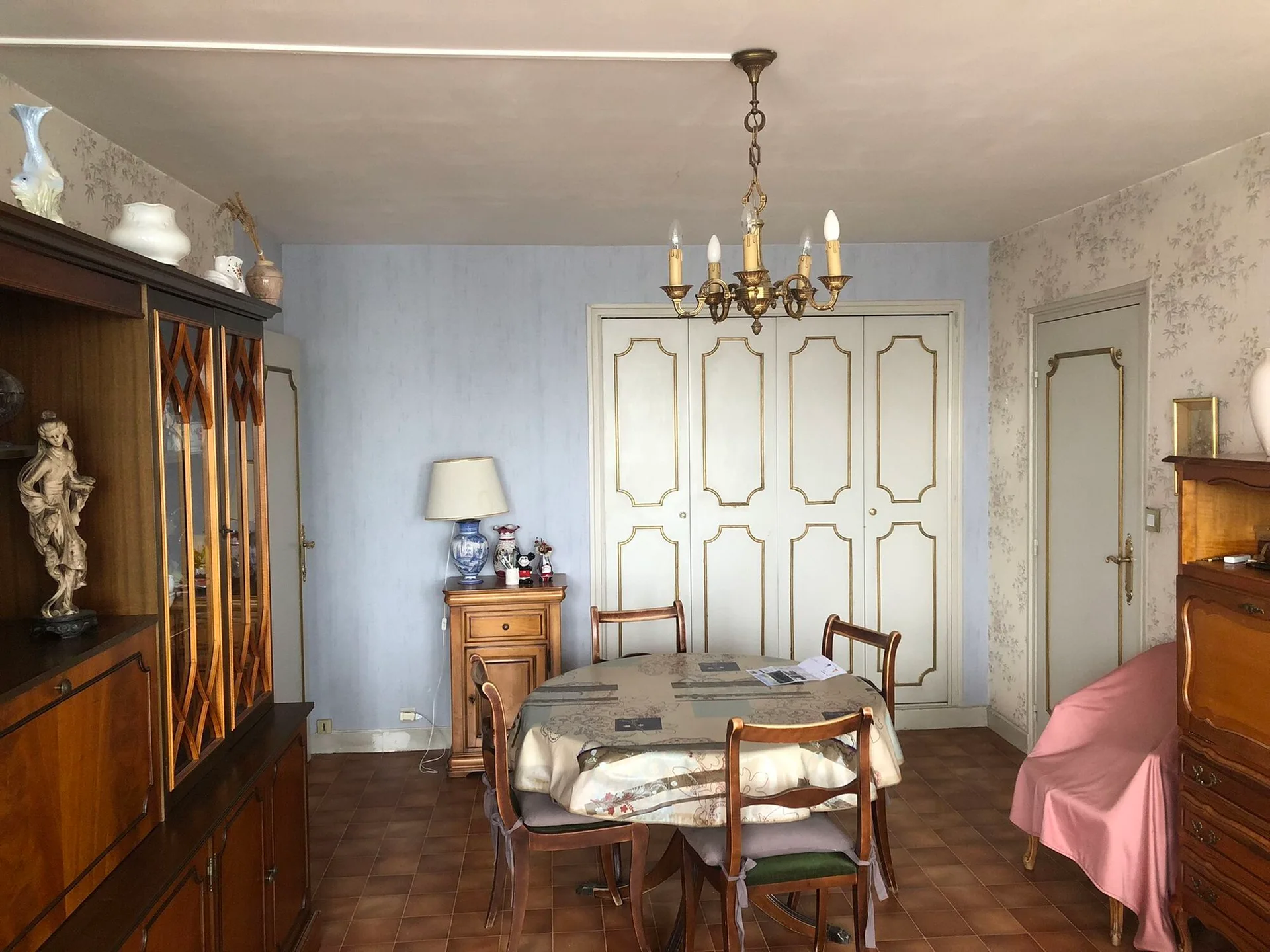 A traditional dining room featuring a round wooden table, white panel closet doors with gold trim, a blue accent wall, and floral wallpaper.
