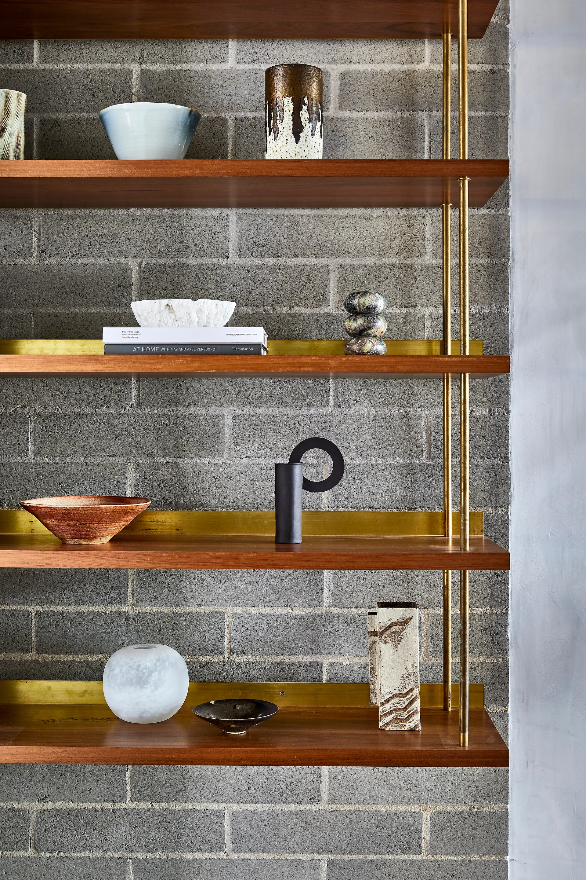A close-up of open wooden shelves supported by slender brass rods, mounted on a gray cinder block wall and styled with ceramic vases.