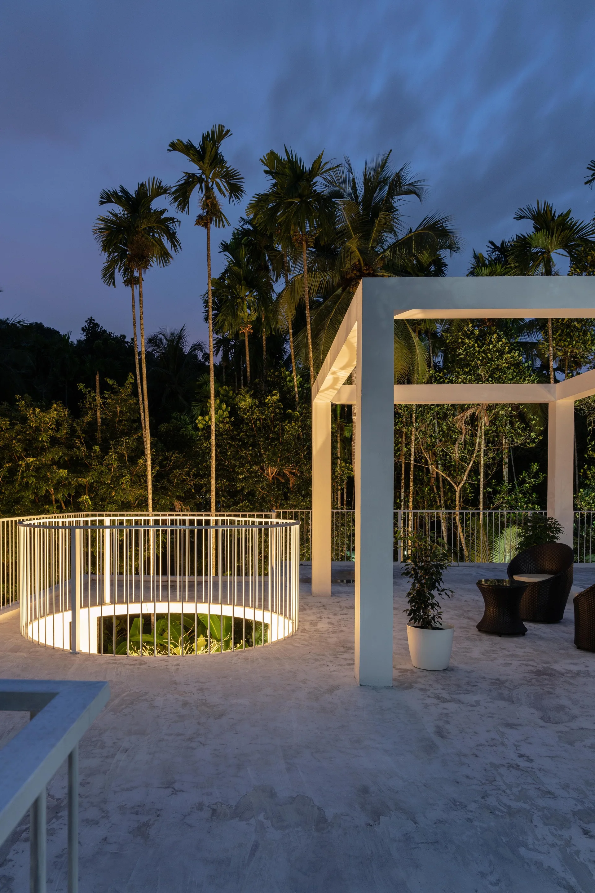 A modern outdoor terrace at twilight features a white geometric canopy and an illuminated circular metal railing surrounding an opening with palm trees behind.