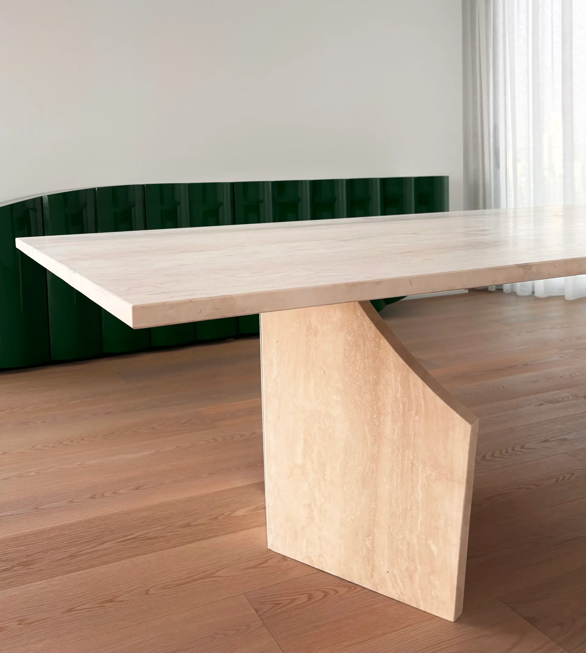 A minimalist beige stone dining table with a geometric pedestal leg stands on light wood floors in front of a low glossy green storage cabinet.