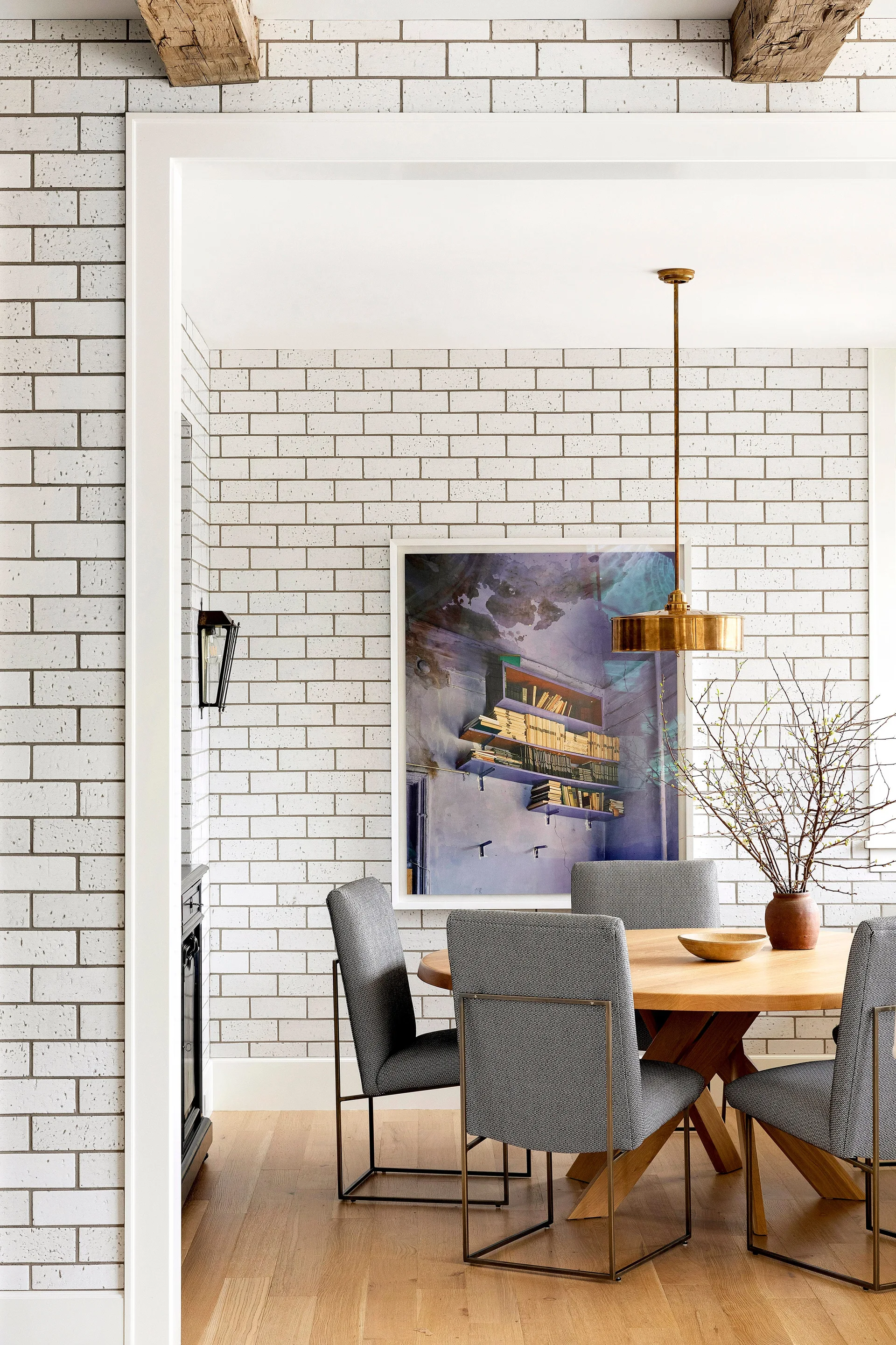 A dining room features a round wooden table, gray upholstered chairs, a white subway tile wall, and a brass pendant light.