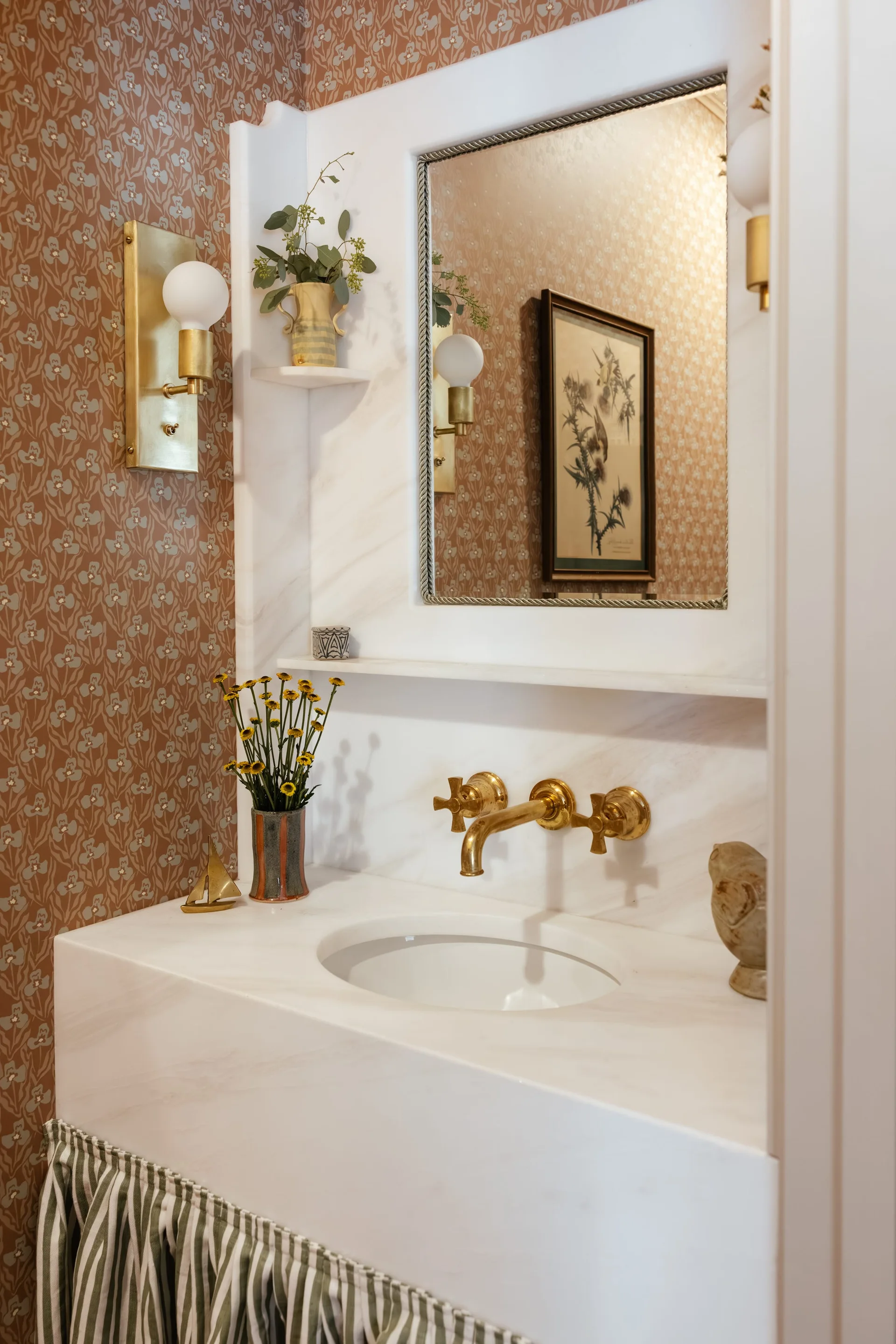 A white marble powder room vanity with a wall-mounted brass faucet, matching mirror, brass sconces, and terracotta floral wallpaper.
