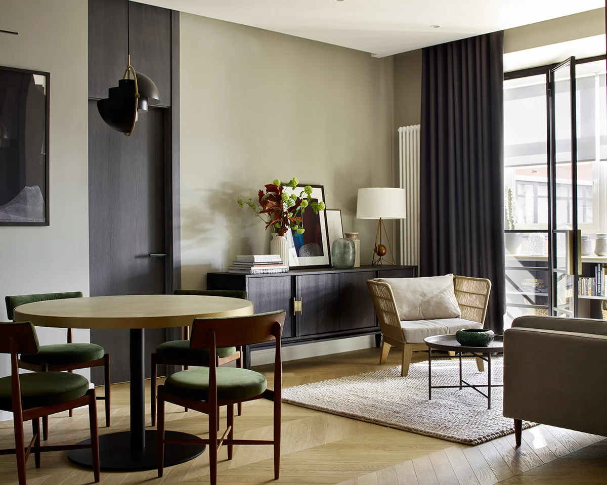 A dining area featuring a round table with green velvet chairs next to a living space with chevron flooring and a dark wood credenza.