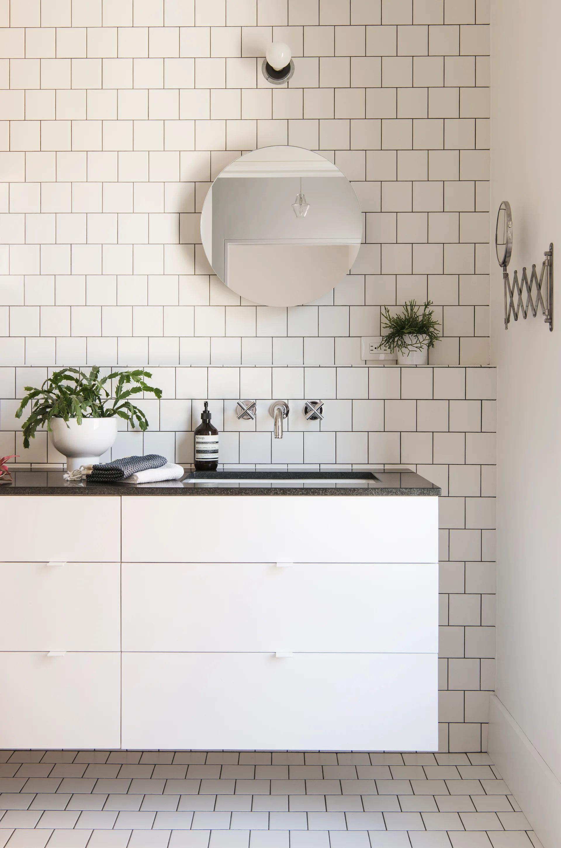 A modern bathroom features a white floating vanity with a black countertop, a round mirror, and white square grid wall and floor tiles.