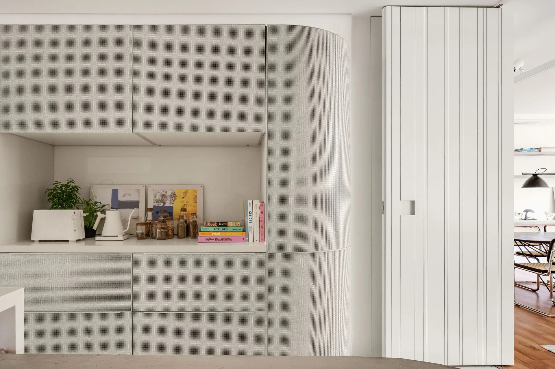 Light gray perforated kitchen cabinetry with a curved corner sits adjacent to a white slatted sliding door and a recessed countertop niche.