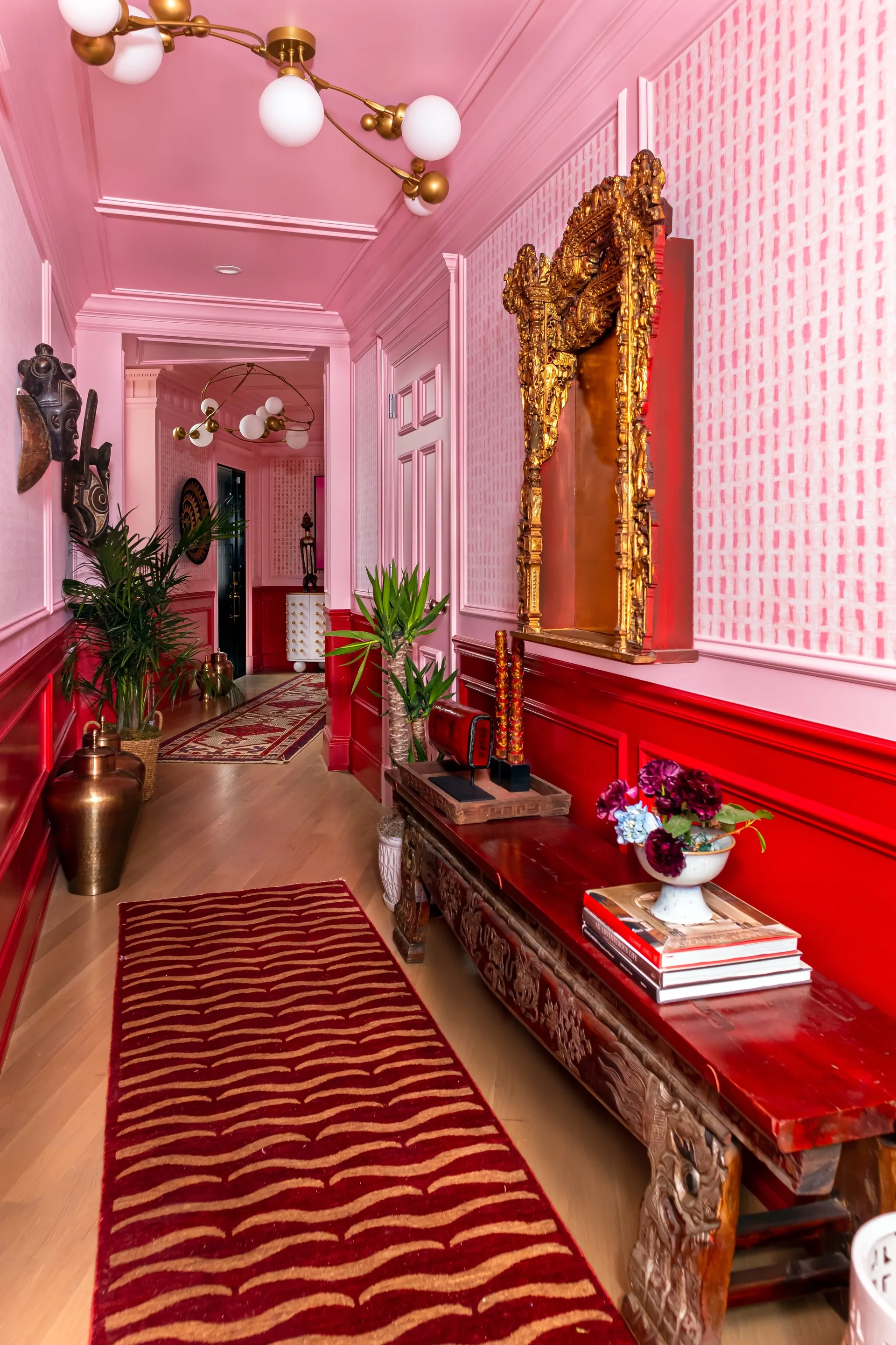 A residential hallway features pink patterned wallpaper, bright red wainscoting, an ornate wooden console table, and a large gold-framed mirror.
