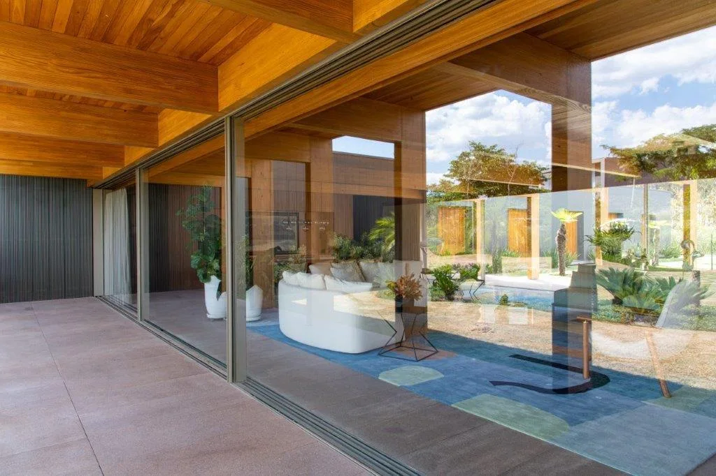 A modern living room viewed through a large sliding glass door, featuring a curved white sofa, geometric blue rug, and an exposed wood ceiling.