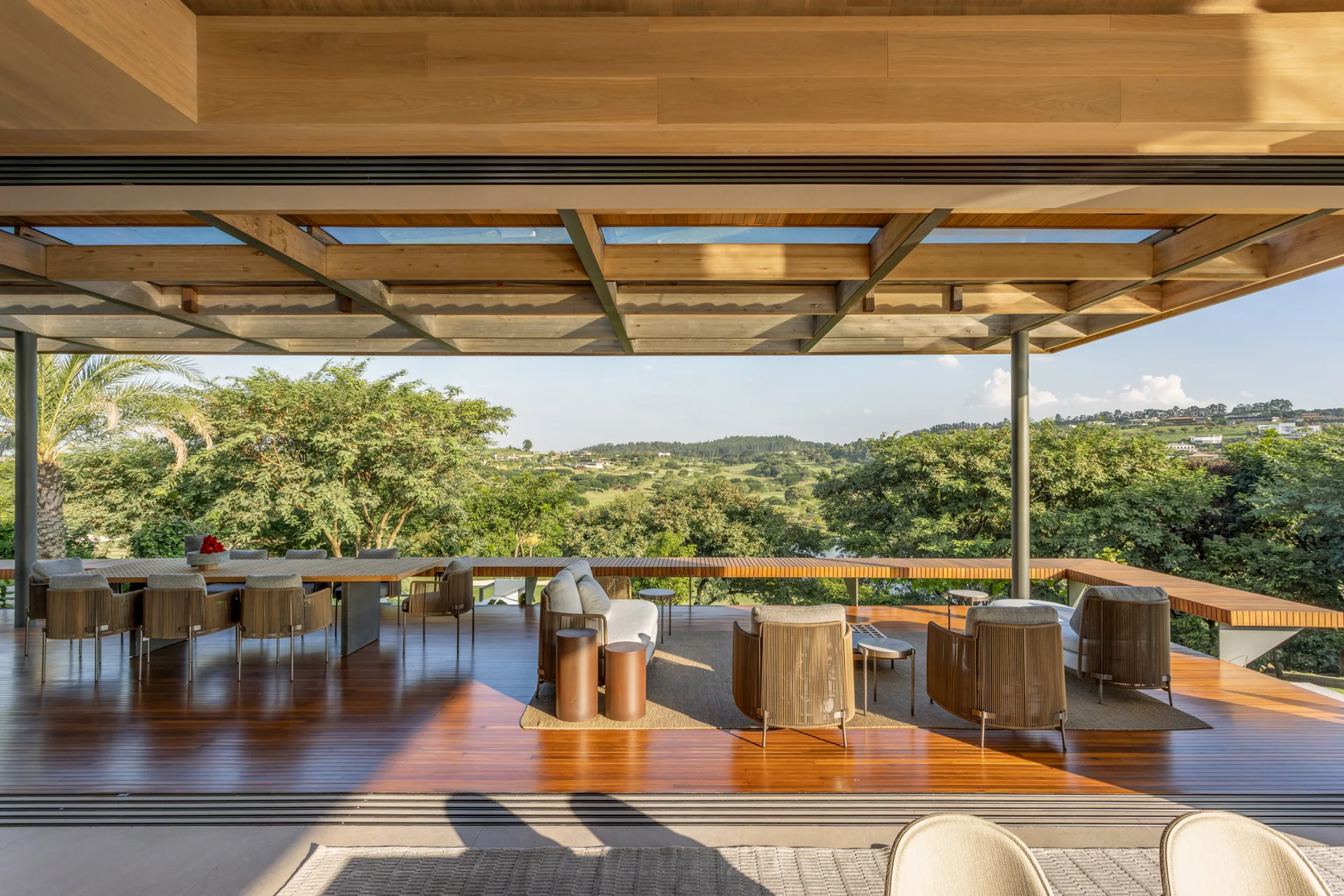 A spacious wood-planked outdoor terrace features a dining table with chairs and a separate lounge seating area under an exposed timber roof with skylights.