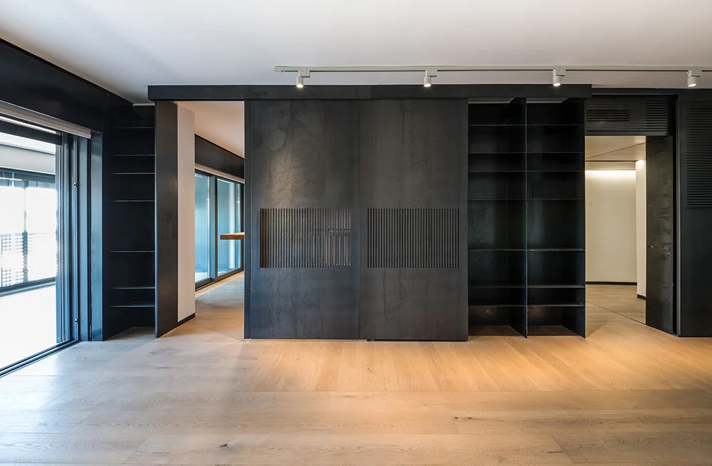 A modern minimalist room featuring a large black metal built-in wall unit with sliding slatted doors, open shelving, and light wood flooring.