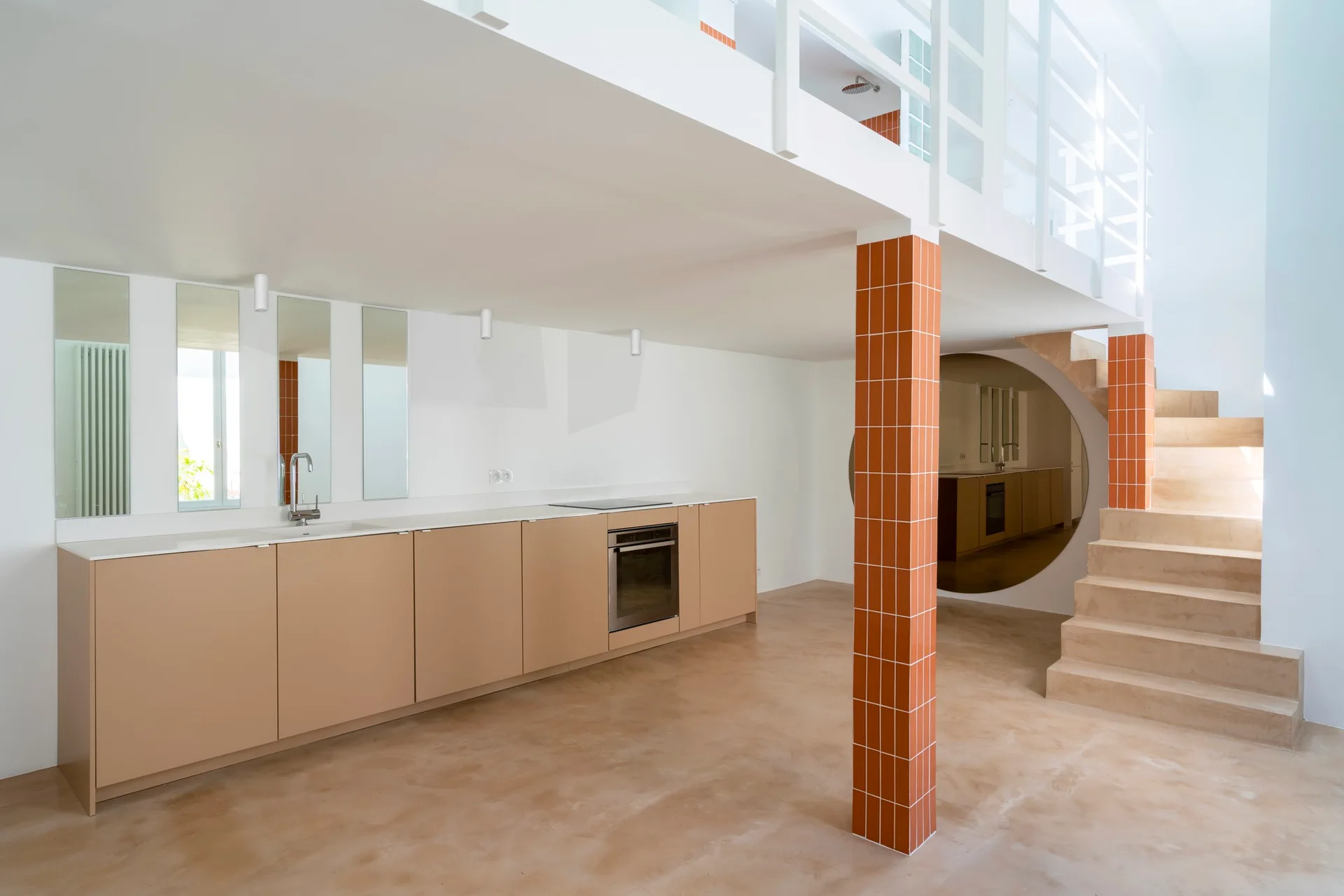 A minimalist kitchen features beige cabinets, an orange tiled column, a concrete-look floor, and a staircase leading to an upper loft.
