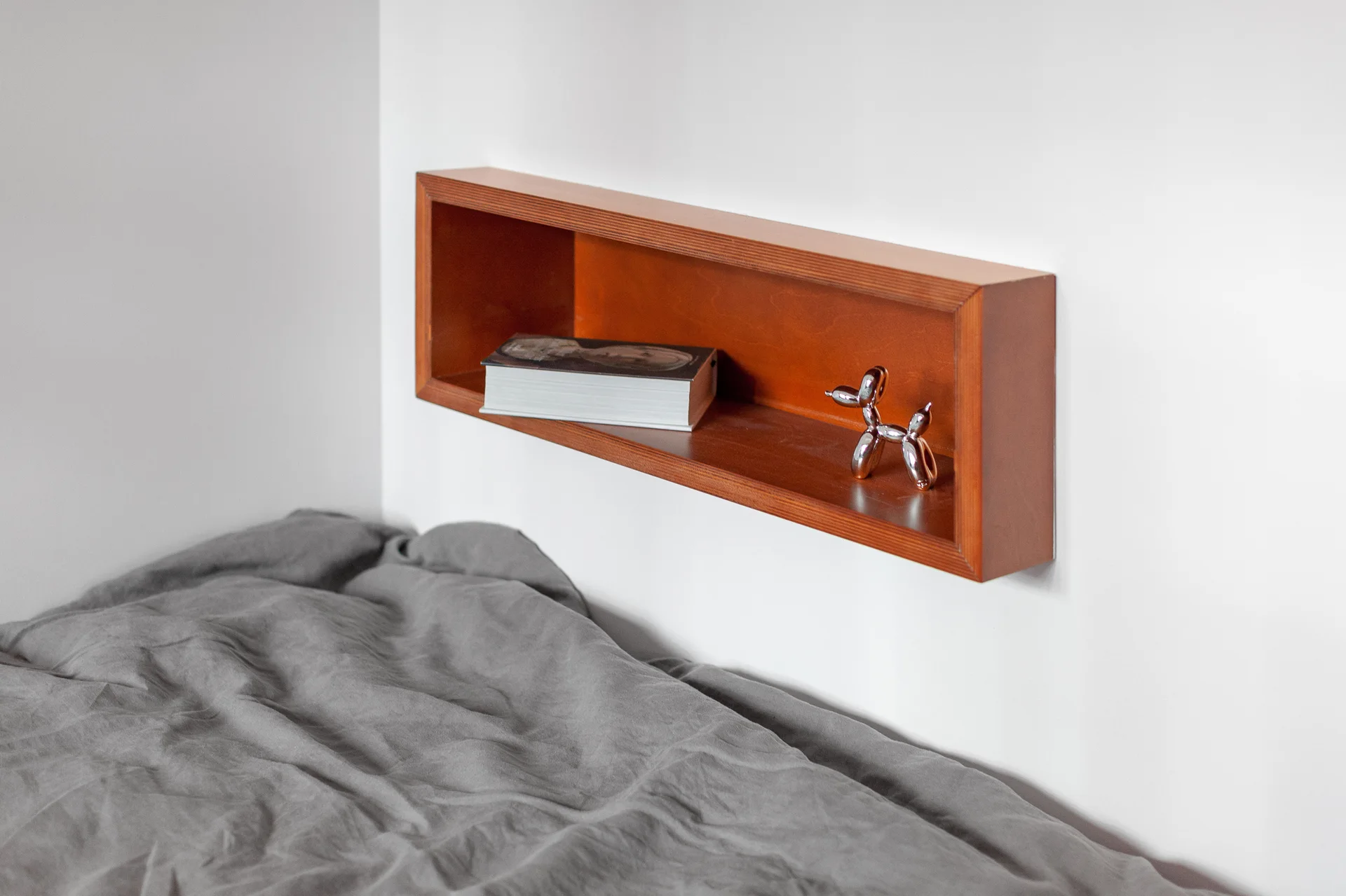 A rectangular wooden shadow box shelf is mounted on a white wall above gray bedding, holding a book and a small metallic balloon dog.