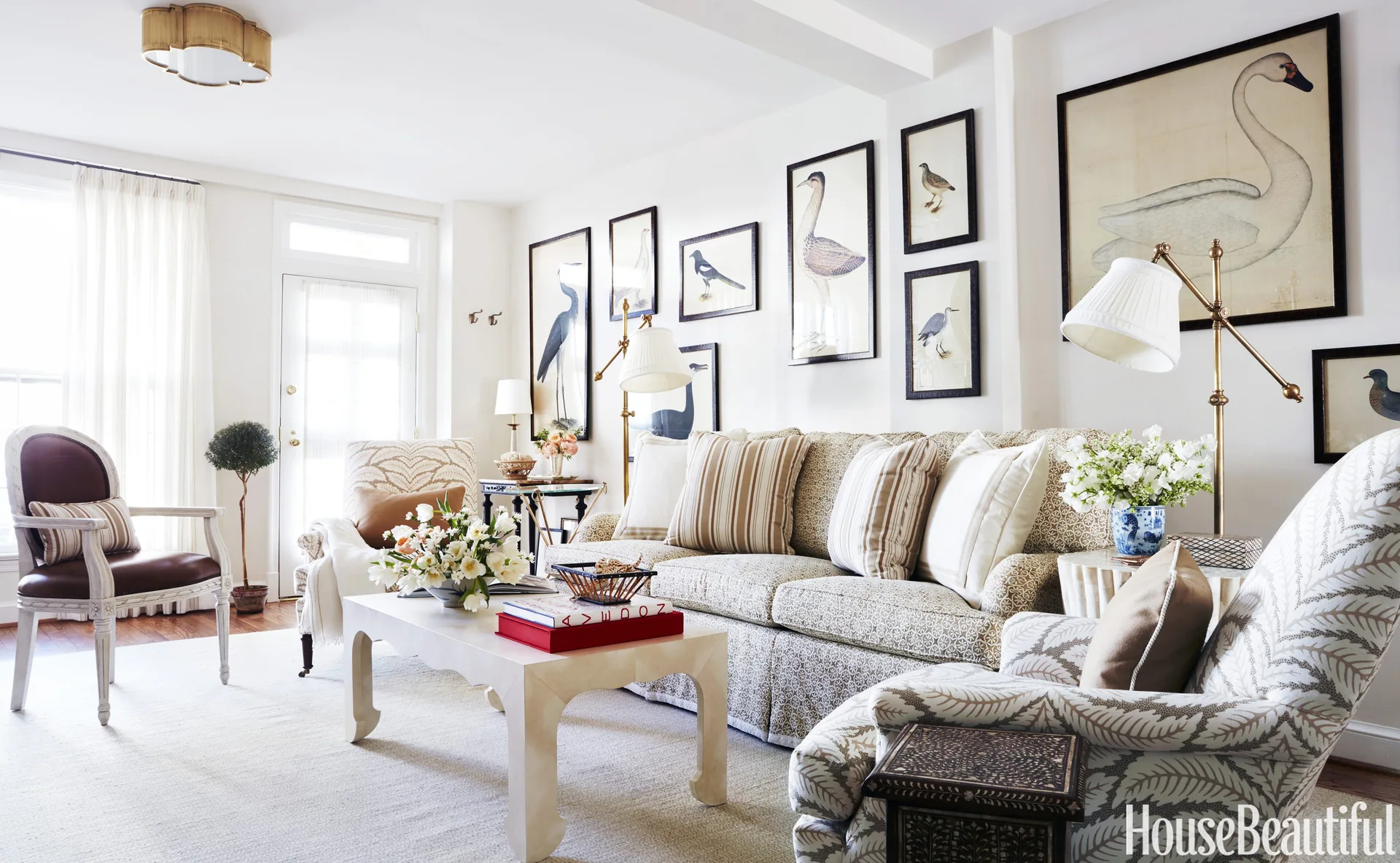A bright living room features a patterned sofa, armchairs, a white coffee table, and a gallery wall of framed bird illustrations.