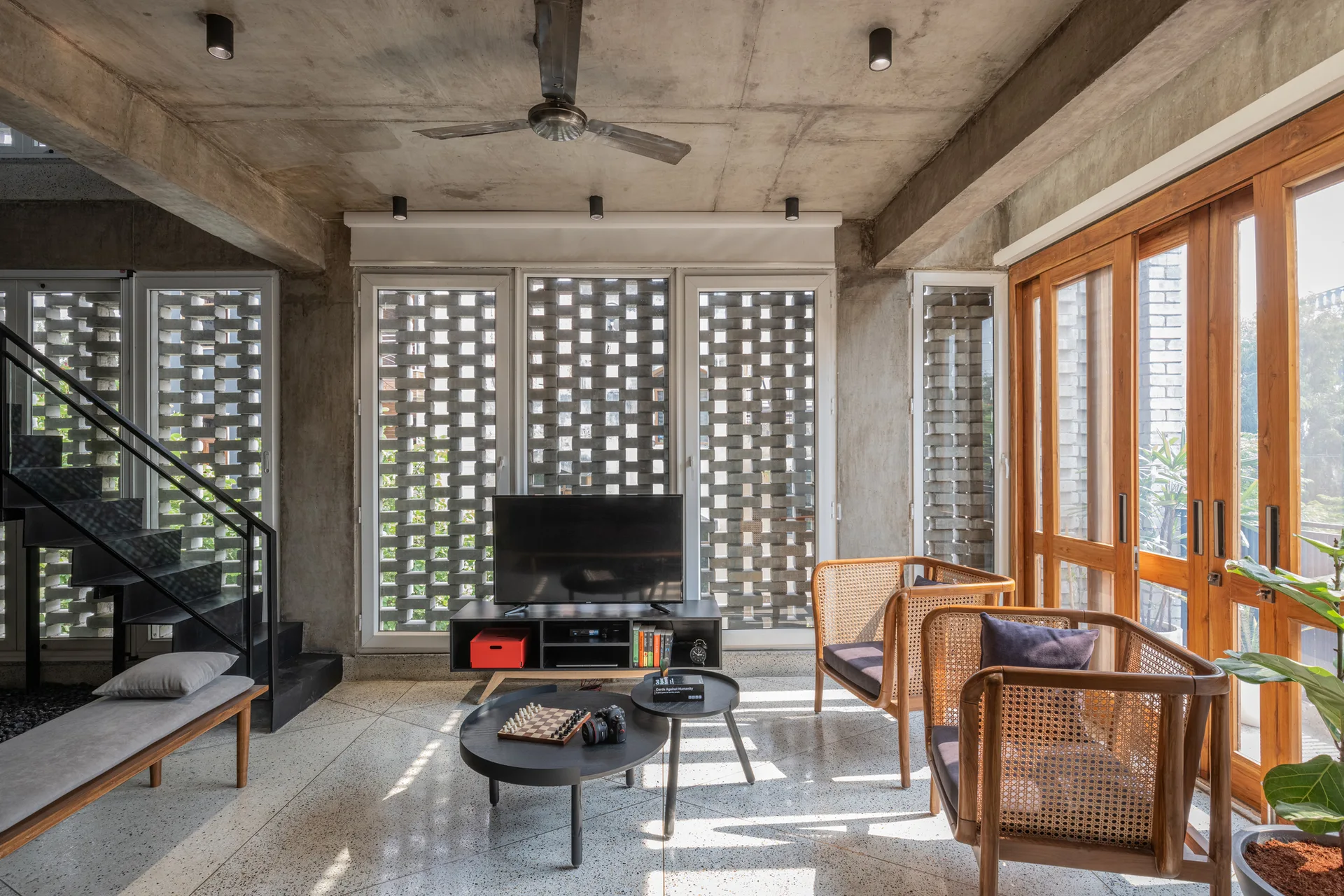 An industrial living room features an exposed concrete ceiling, a perforated breeze block wall, two wooden cane armchairs, and a terrazzo floor.