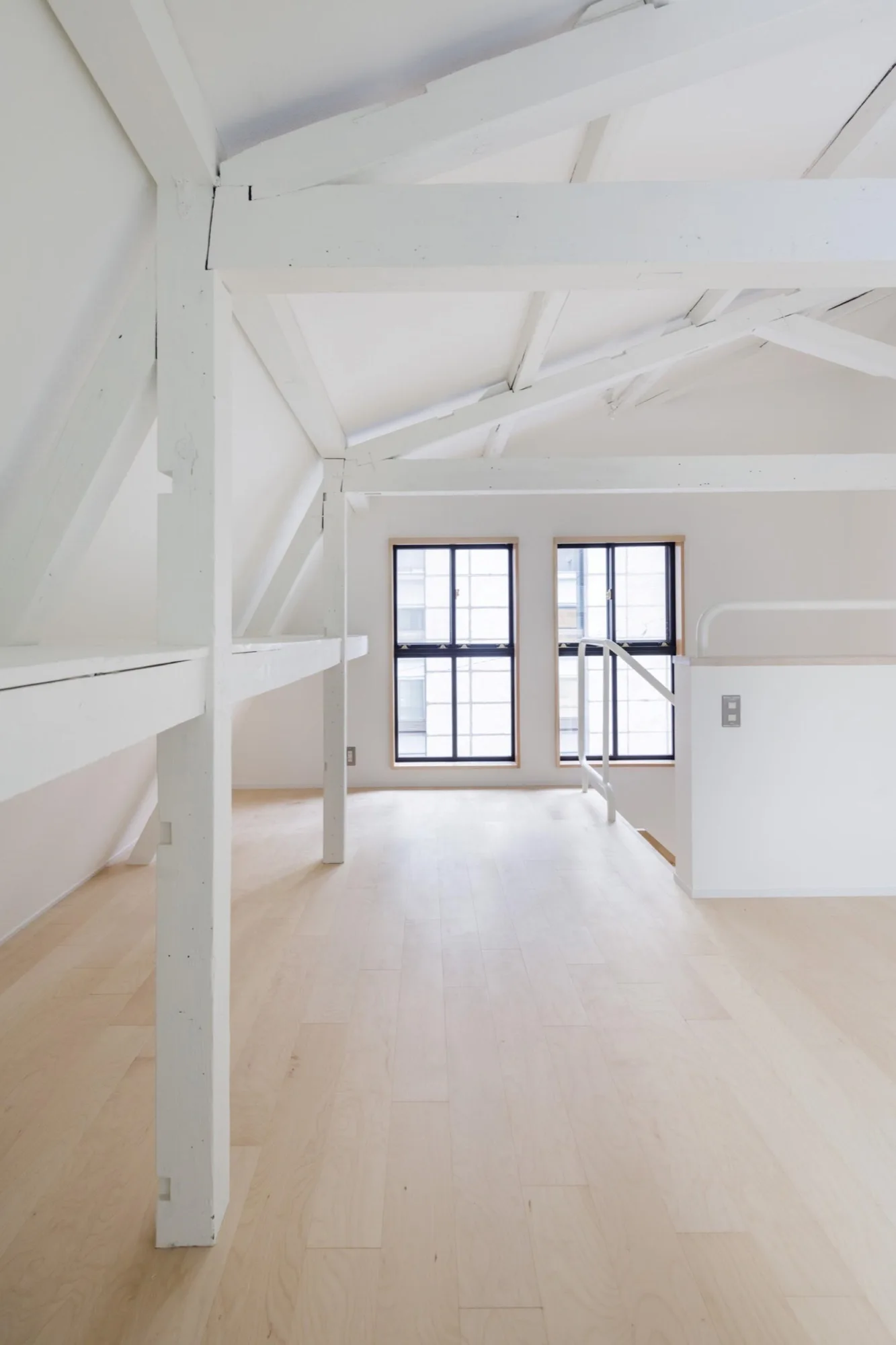 A minimalist attic landing features light blonde wood floors, white painted walls and exposed timber beams, and two black-framed windows.