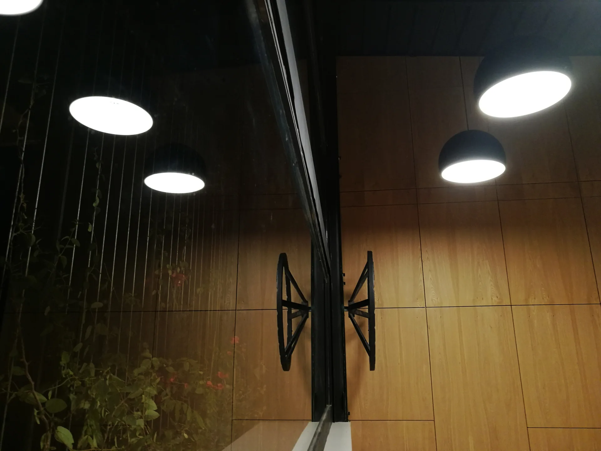 Modern black dome pendant lights hang in front of a warm wood-paneled wall next to a glass window reflecting green foliage outside.