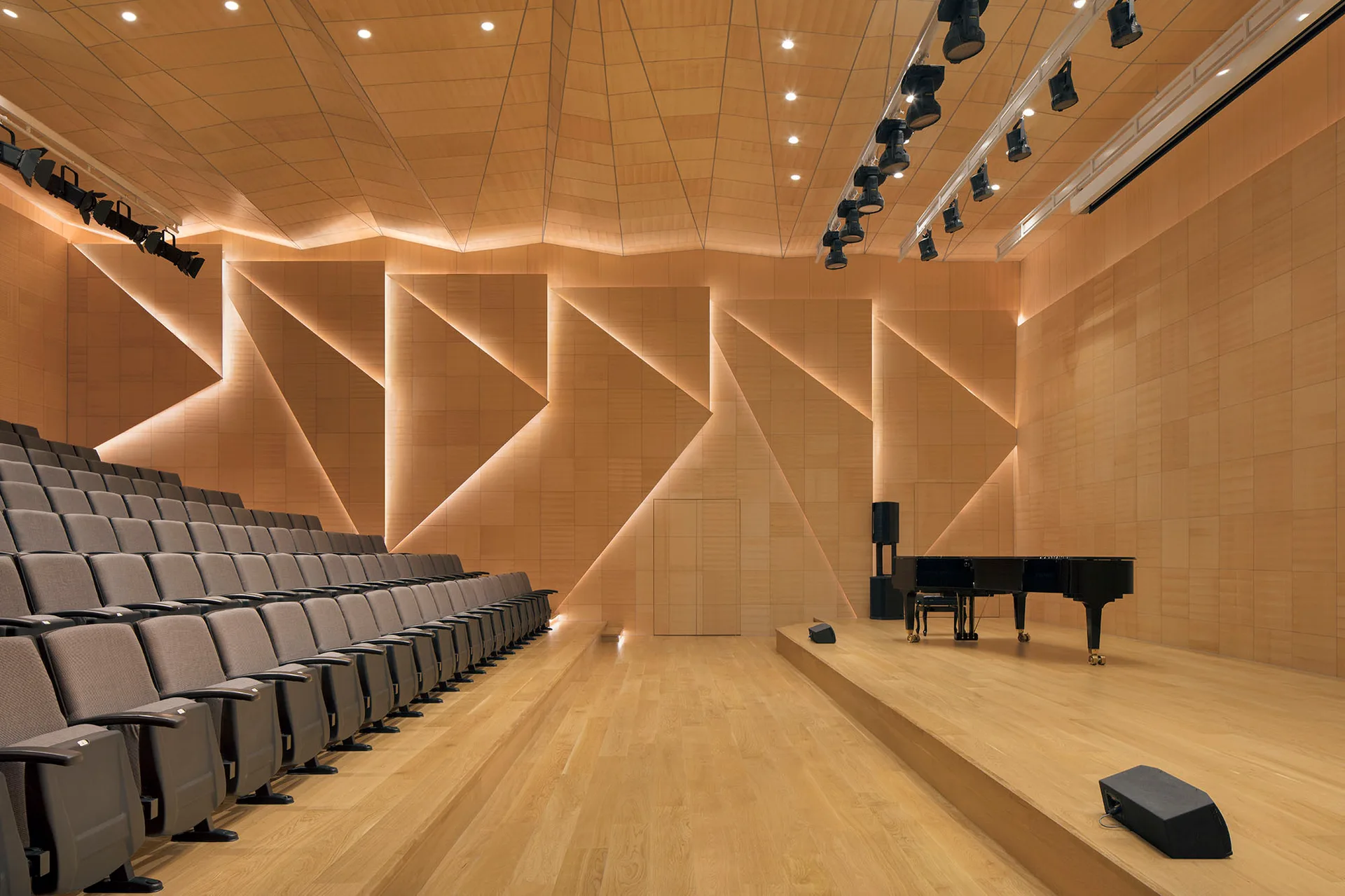 A modern auditorium features angled wood-paneled walls with recessed lighting, tiered grey seating, and a black grand piano on a wooden stage.