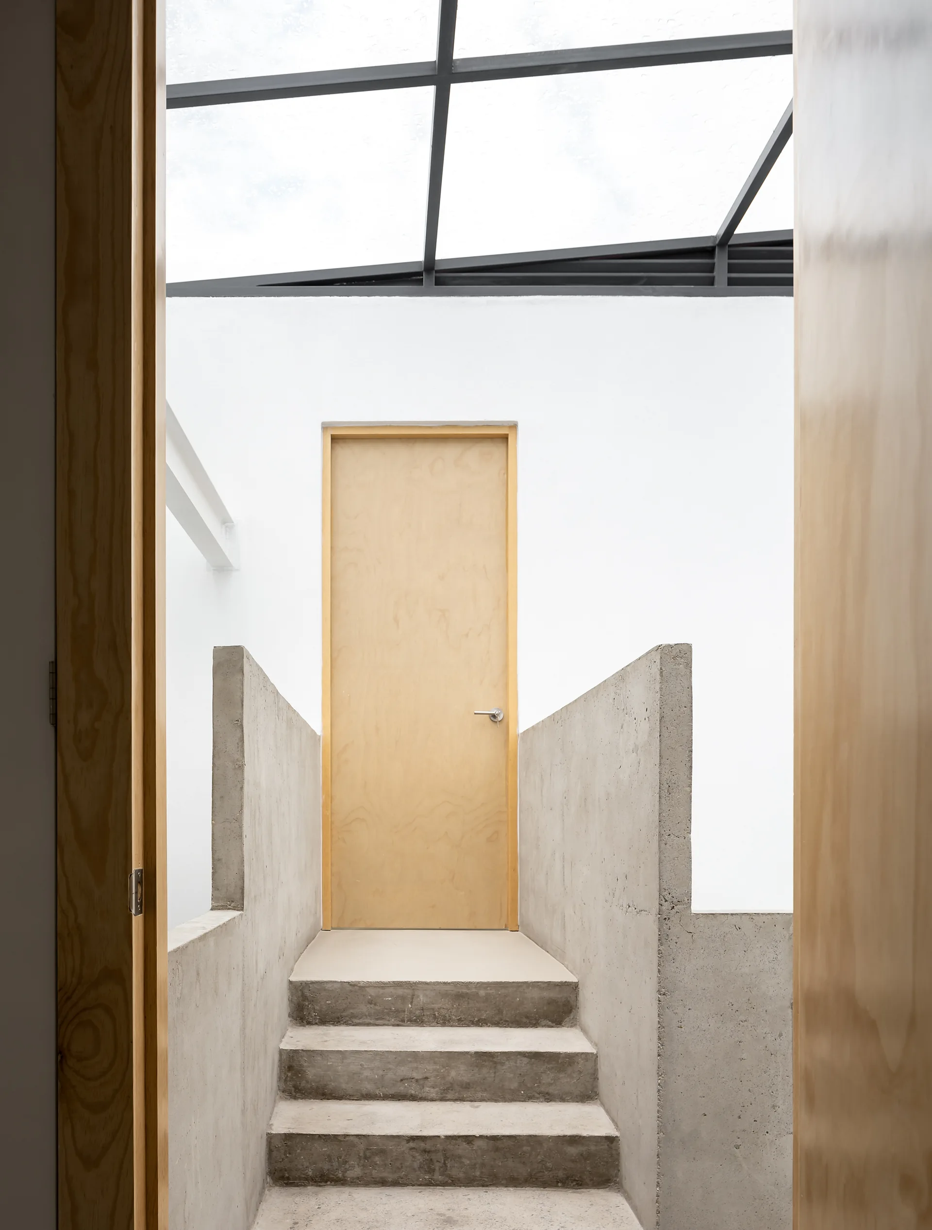 A minimalist concrete staircase with raw concrete walls leads to a light wood door beneath a black metal skylight.