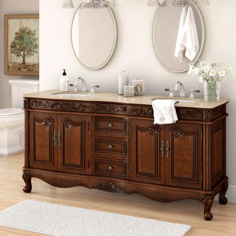 A traditional brown wood double vanity with ornate carvings, a beige stone countertop, and two oval silver-framed mirrors on a light wall.