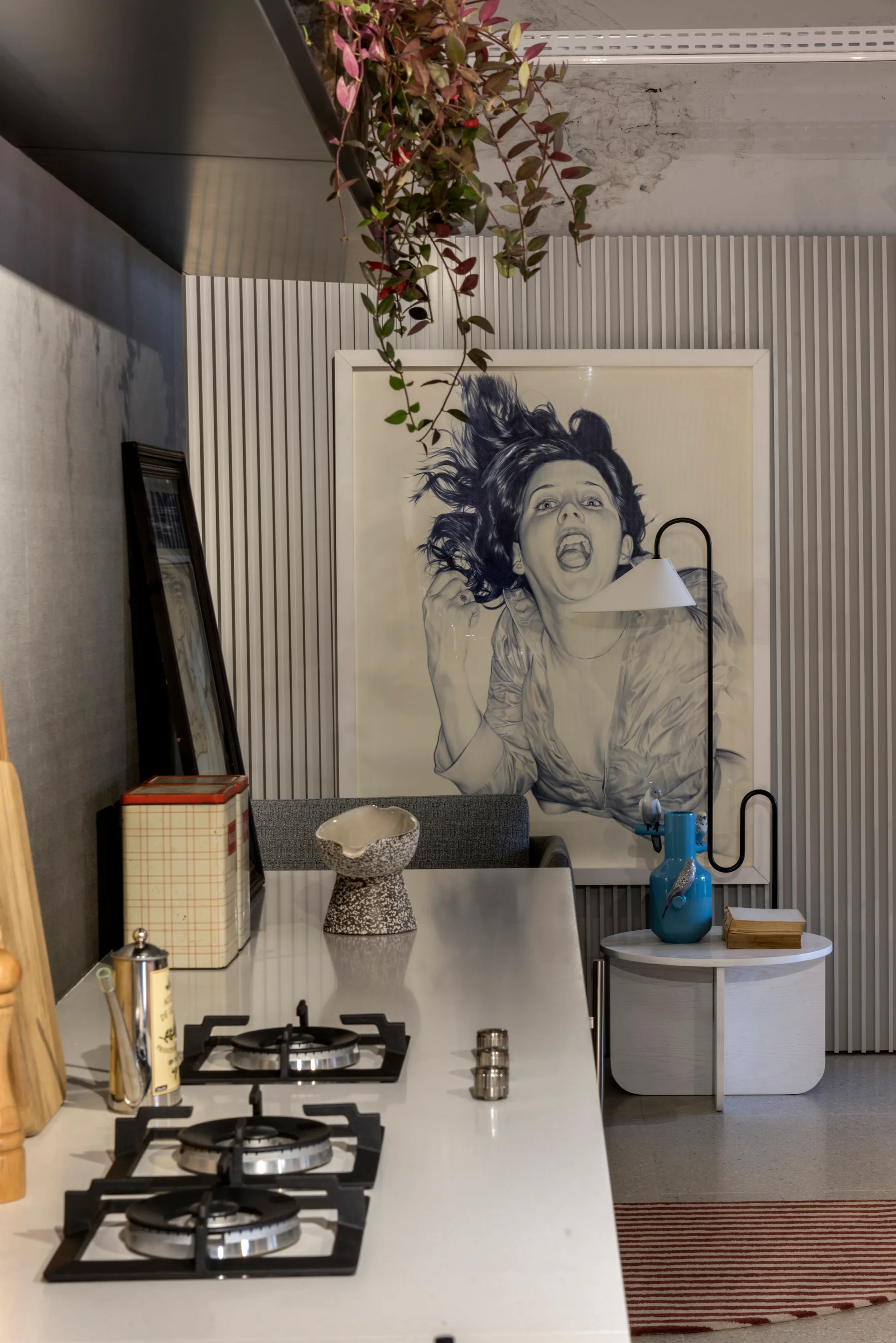 A white kitchen countertop with black gas burners sits before a vertical slatted wall featuring a large framed graphic portrait print.