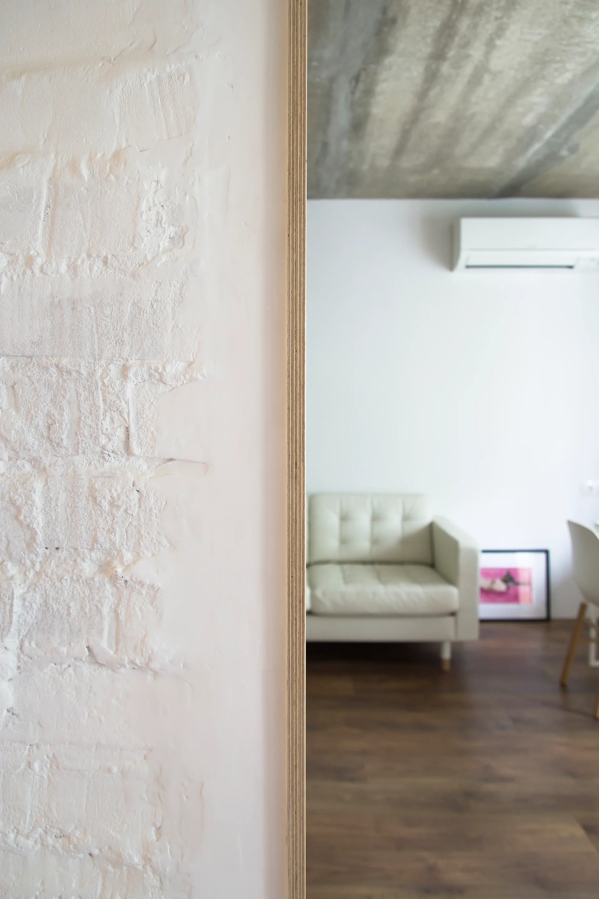 A white painted brick wall with an exposed plywood edge frames a minimalist living room featuring a light tufted sofa and a raw concrete ceiling.
