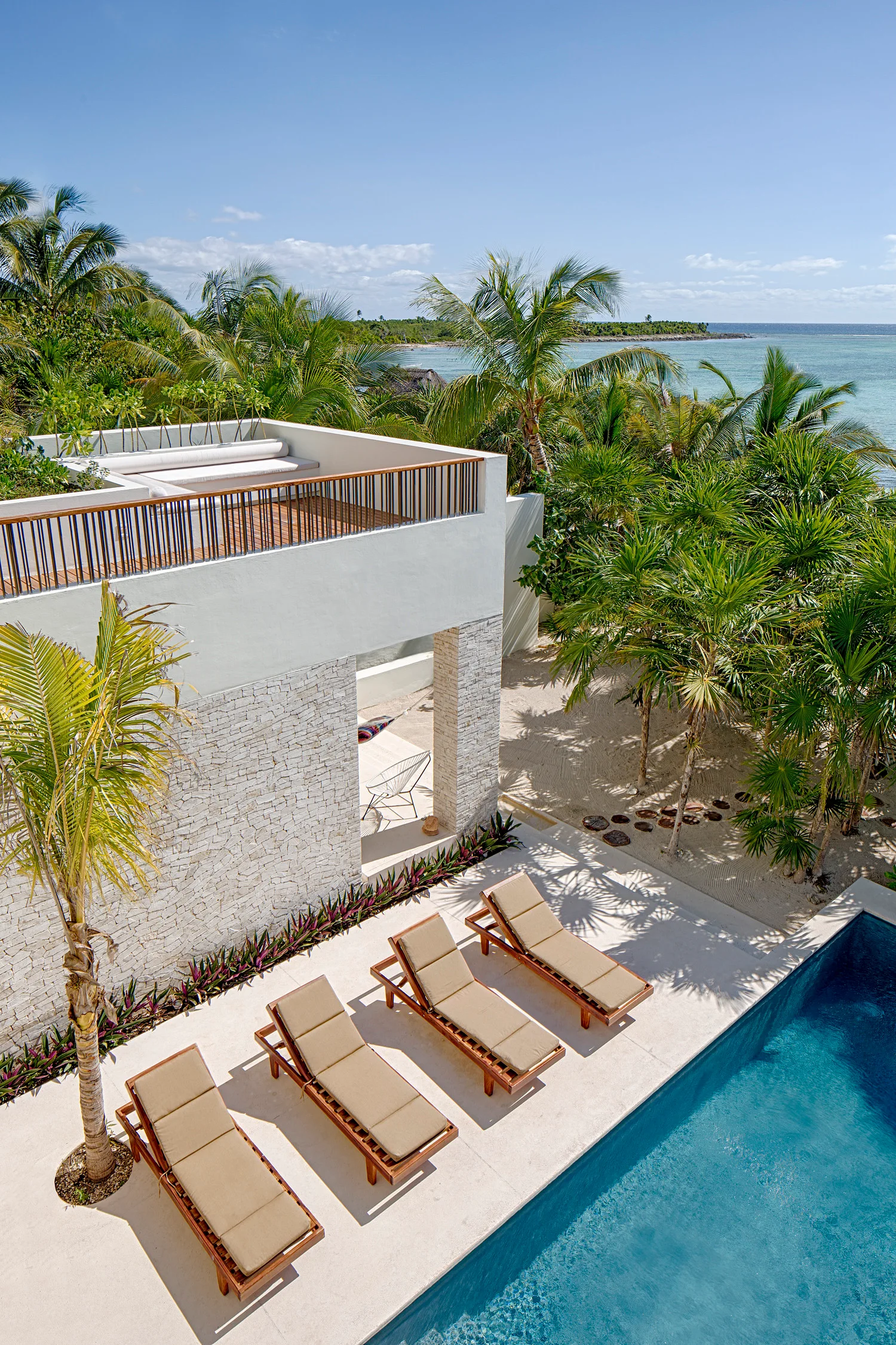A high-angle view of a tropical poolside patio with four tan lounge chairs, a white stone building, and a rooftop terrace overlooking the ocean.