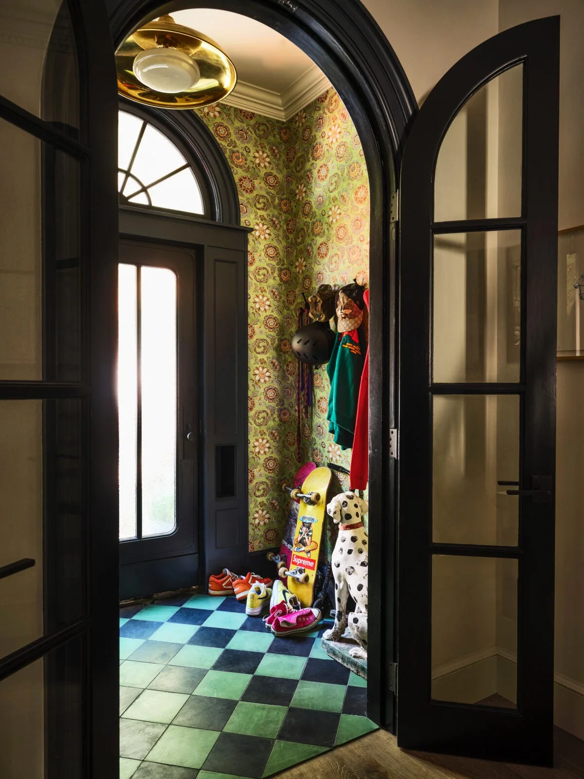 A view through arched black doors into a maximalist entryway featuring green floral wallpaper and a black and green checkered tile floor.