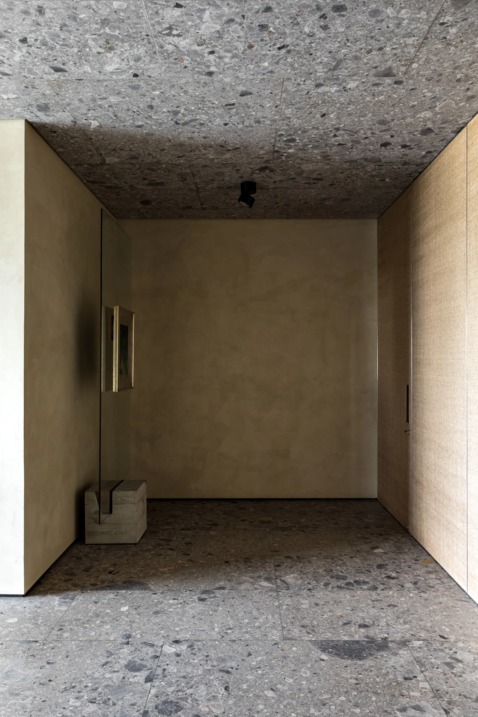 A minimalist corridor features matching grey terrazzo floors and ceilings, textured plaster walls, and a glass partition displaying a framed artwork.