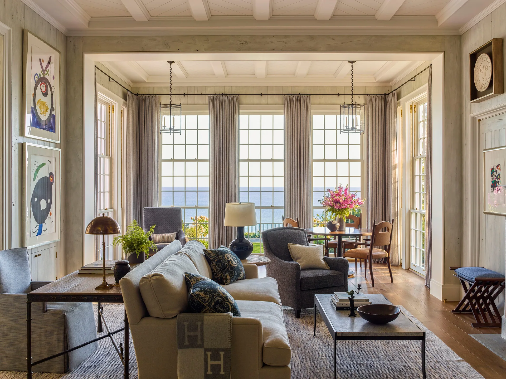 A coastal living room features a light grey sofa, dark armchairs, and a small dining table in front of large windows with an ocean view.