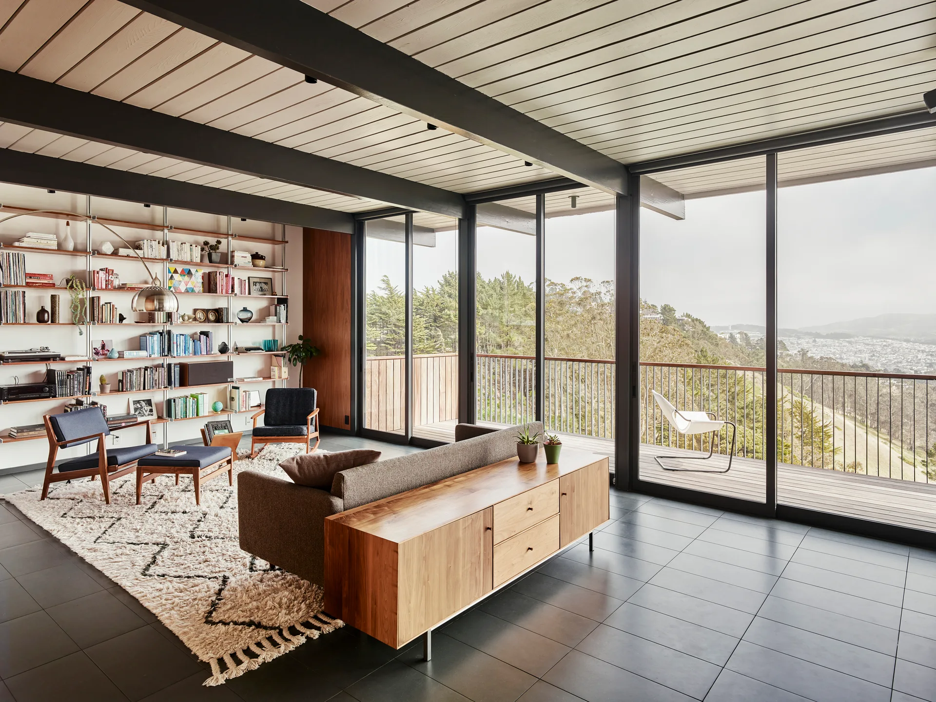 A mid-century living room features a wall-mounted shelving unit, a gray sofa backed by a wooden credenza, and floor-to-ceiling glass sliding doors.