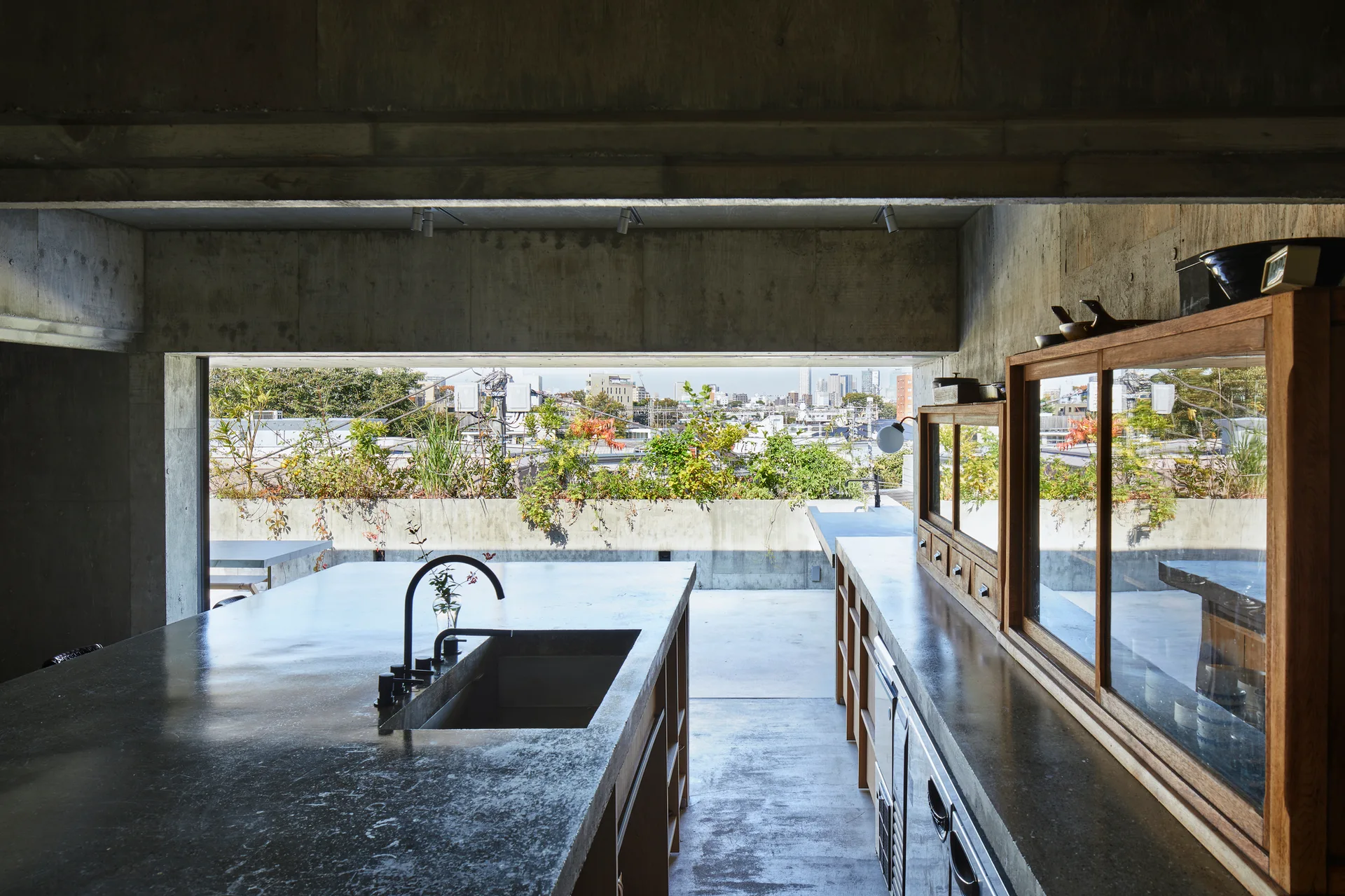 A minimalist kitchen features a raw concrete island with a black faucet, wooden cabinets, and a wide horizontal window opening to an outdoor terrace.