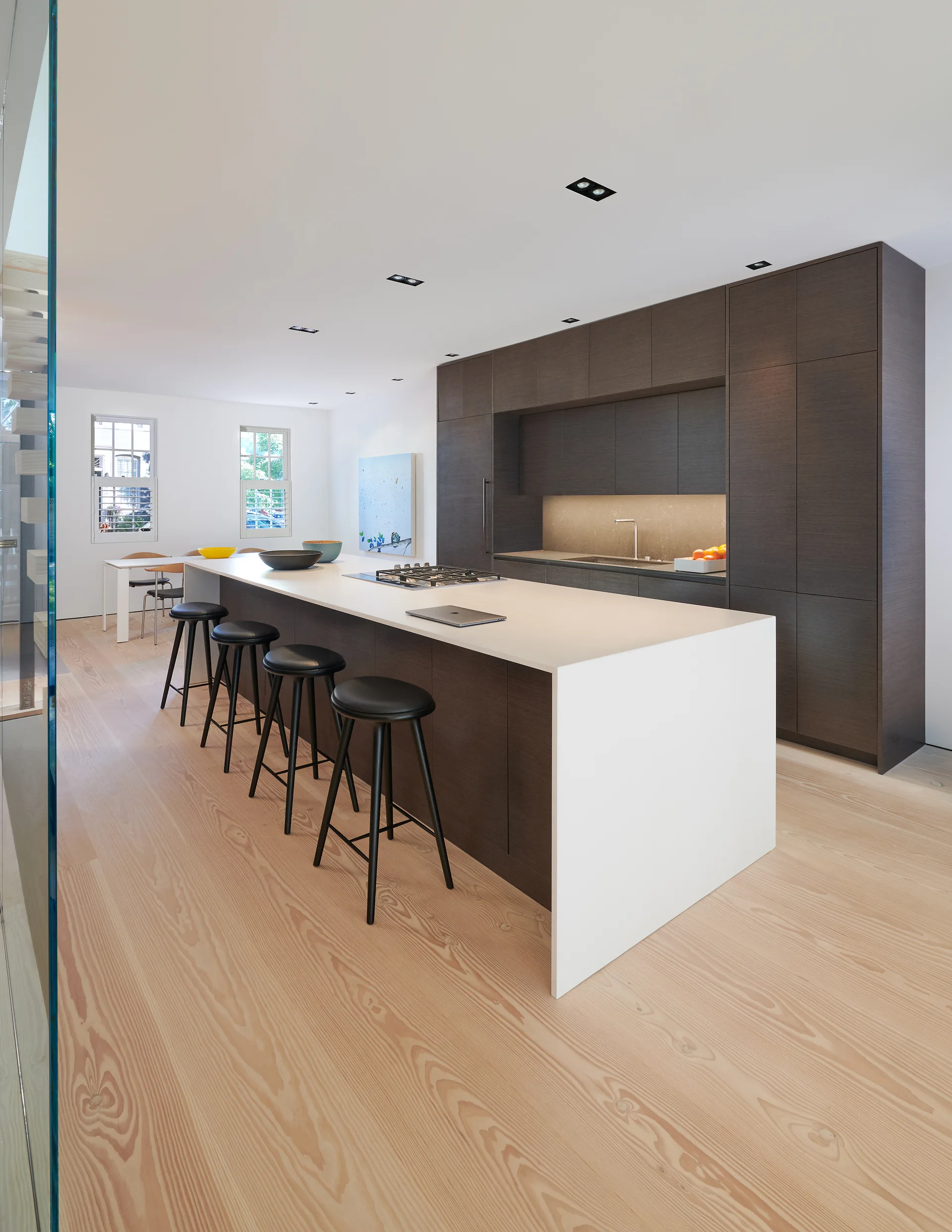 A modern kitchen features a long white waterfall island with black bar stools, dark wood cabinetry, and light wood flooring.