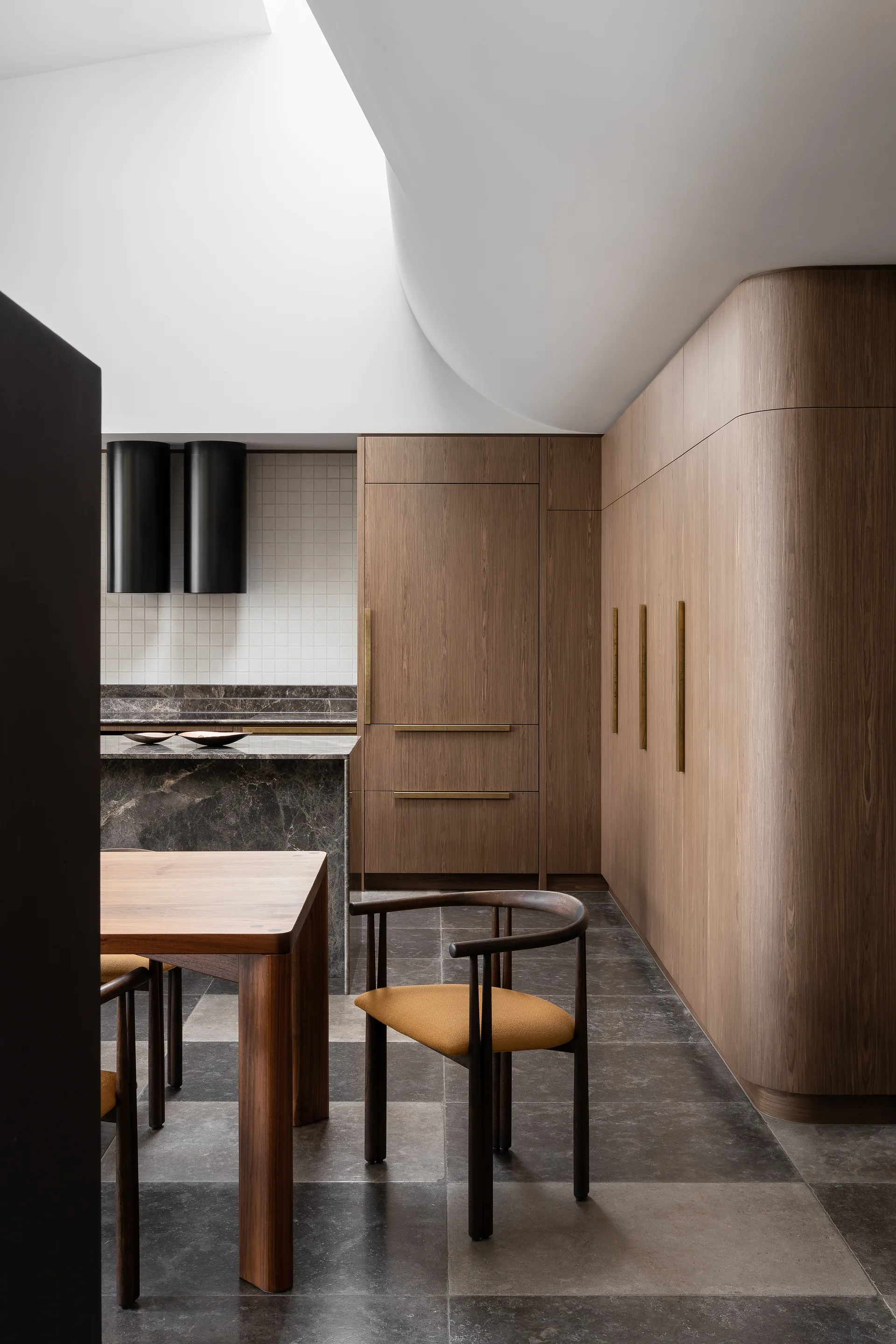 A contemporary kitchen features curved wood cabinetry, a dark marble island, and a dining chair with a mustard cushion under a curved white ceiling.