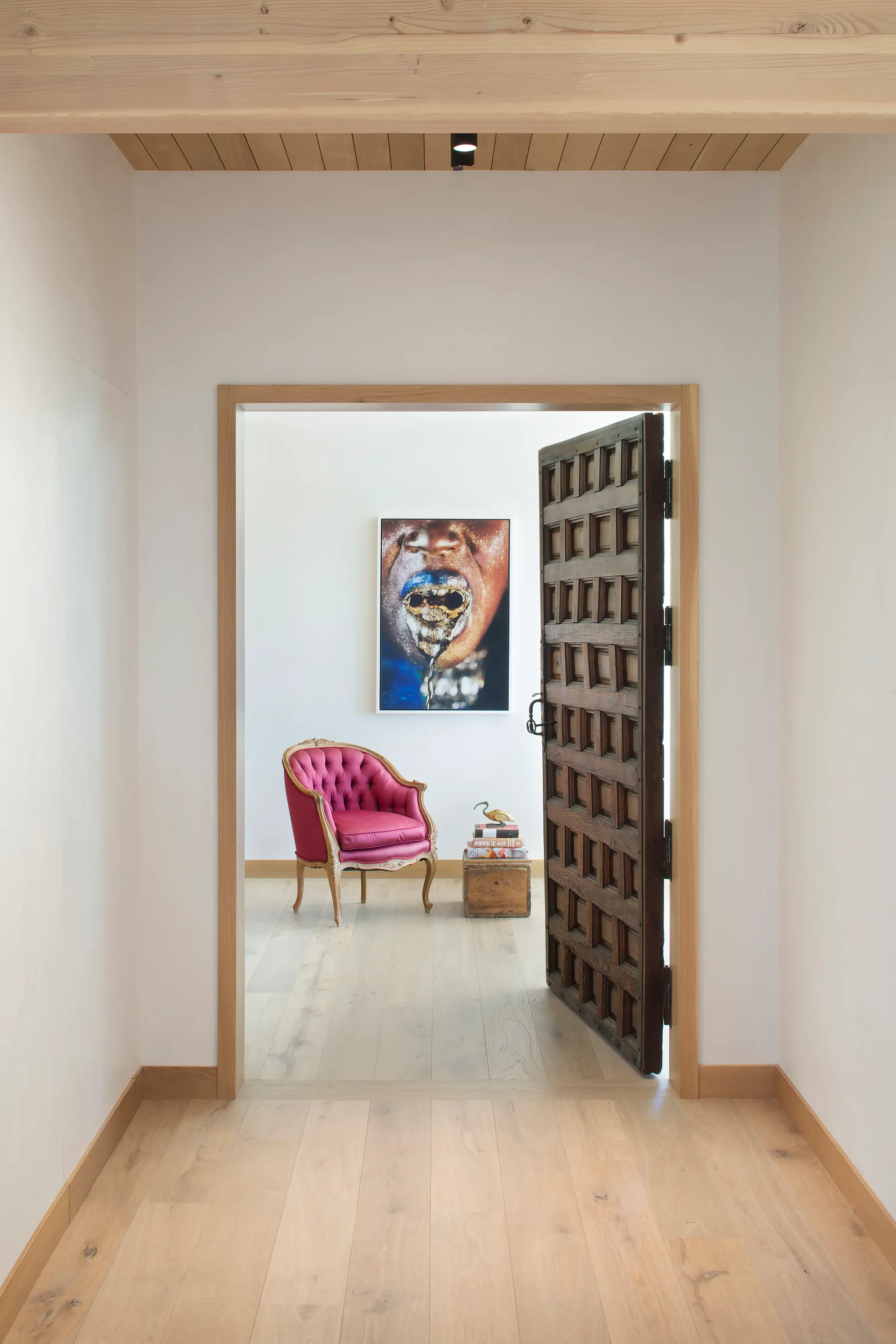 A view through an open carved wood door reveals a white room with a tufted magenta armchair, framed artwork, and light wood flooring.