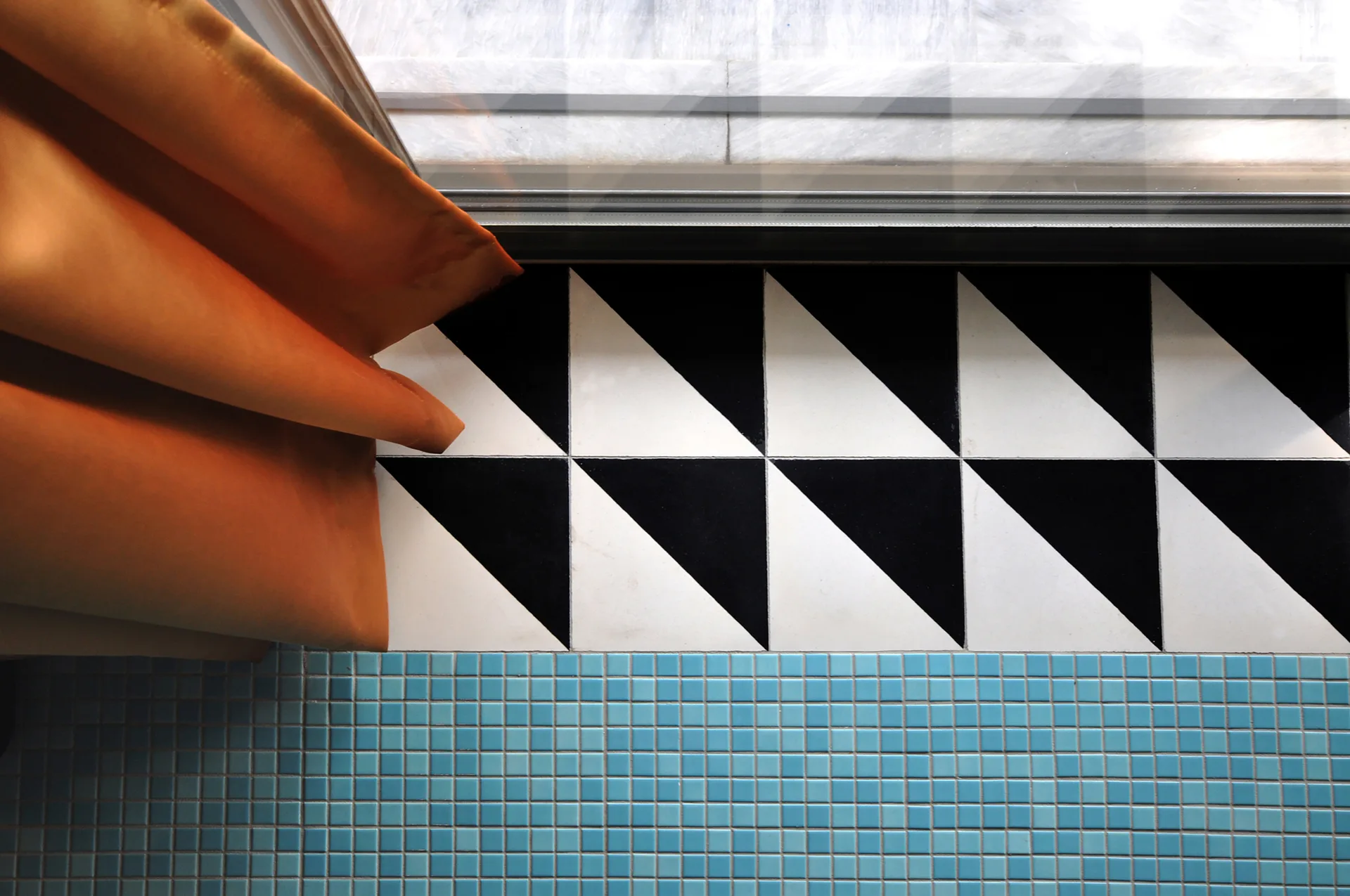 A top-down view of a floor transition from black and white triangular tiles to small light blue mosaic tiles, with an orange curtain on the left.