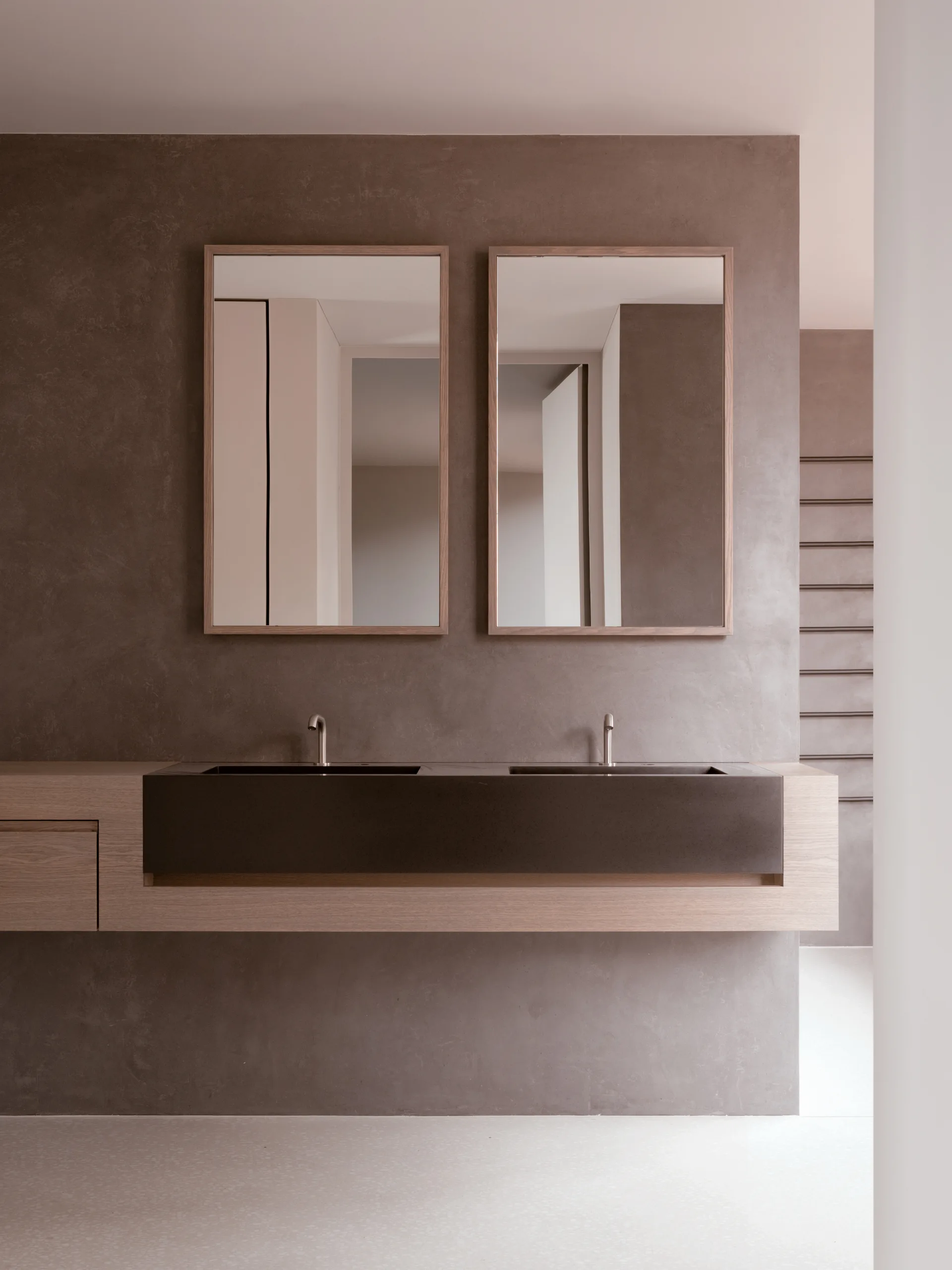 A floating wood bathroom vanity with a dark inset double sink sits beneath two rectangular wood-framed mirrors against a textured taupe wall.