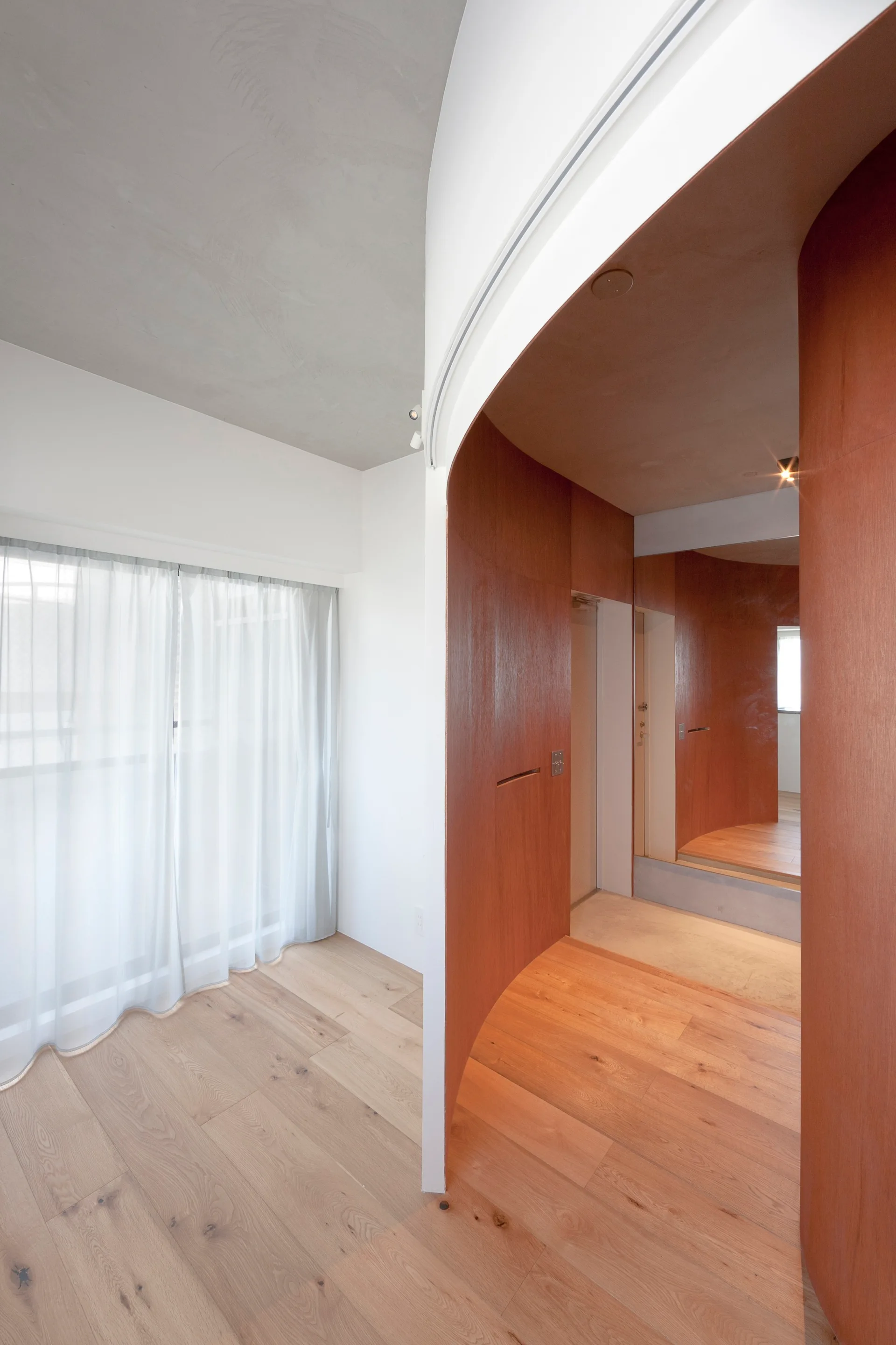 A curved wood partition wall stands on light oak flooring next to sheer white window curtains and a concrete ceiling.