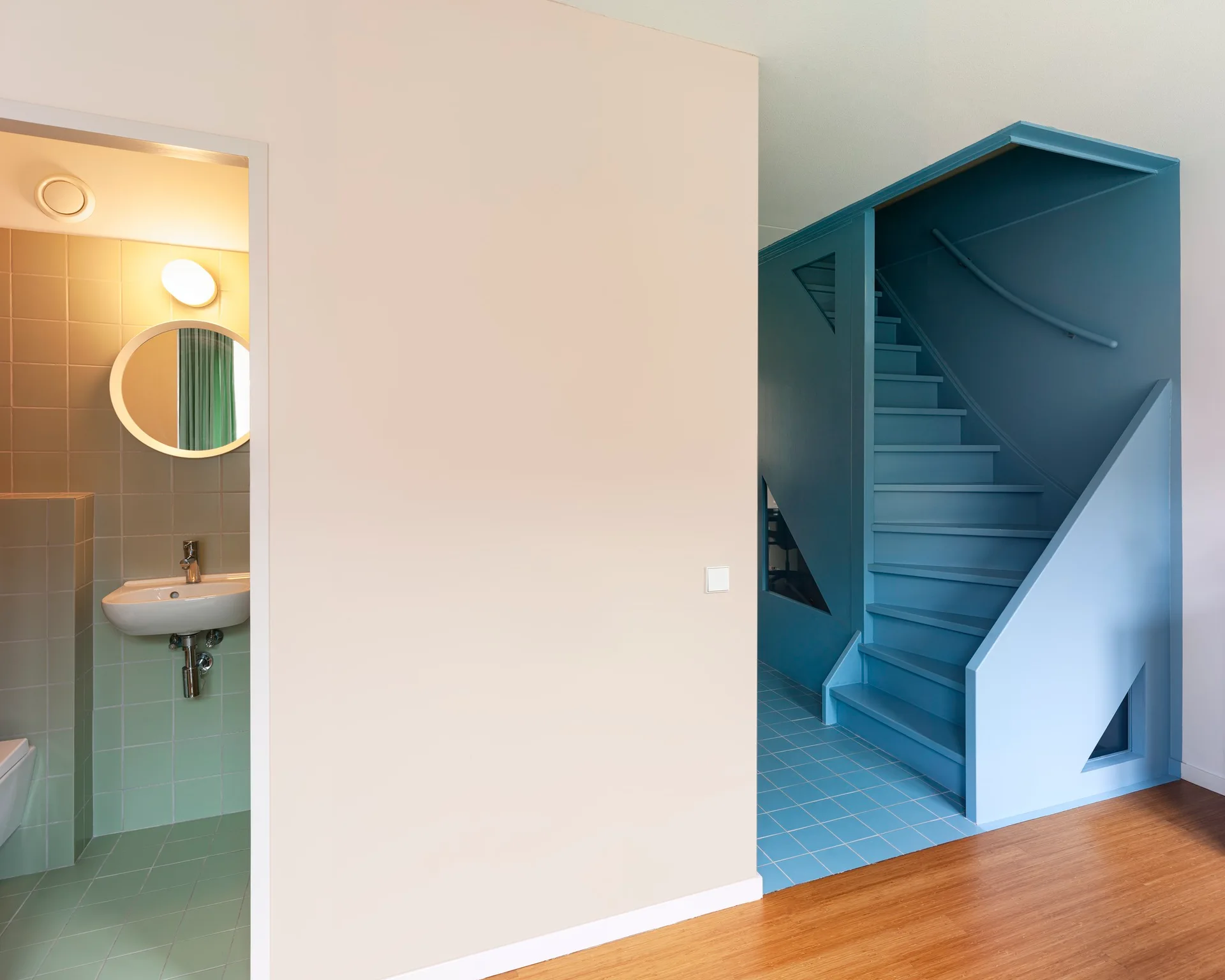 A beige partition wall stands between a green tiled bathroom with a round mirror and a bold blue staircase on matching blue tiles.