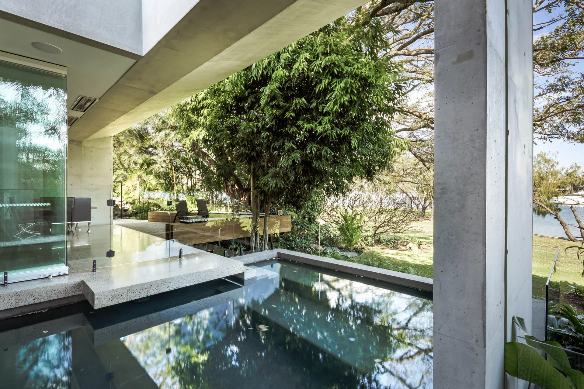 A modern concrete patio and swimming pool with transparent glass railings overlooking a lush green garden with mature trees.