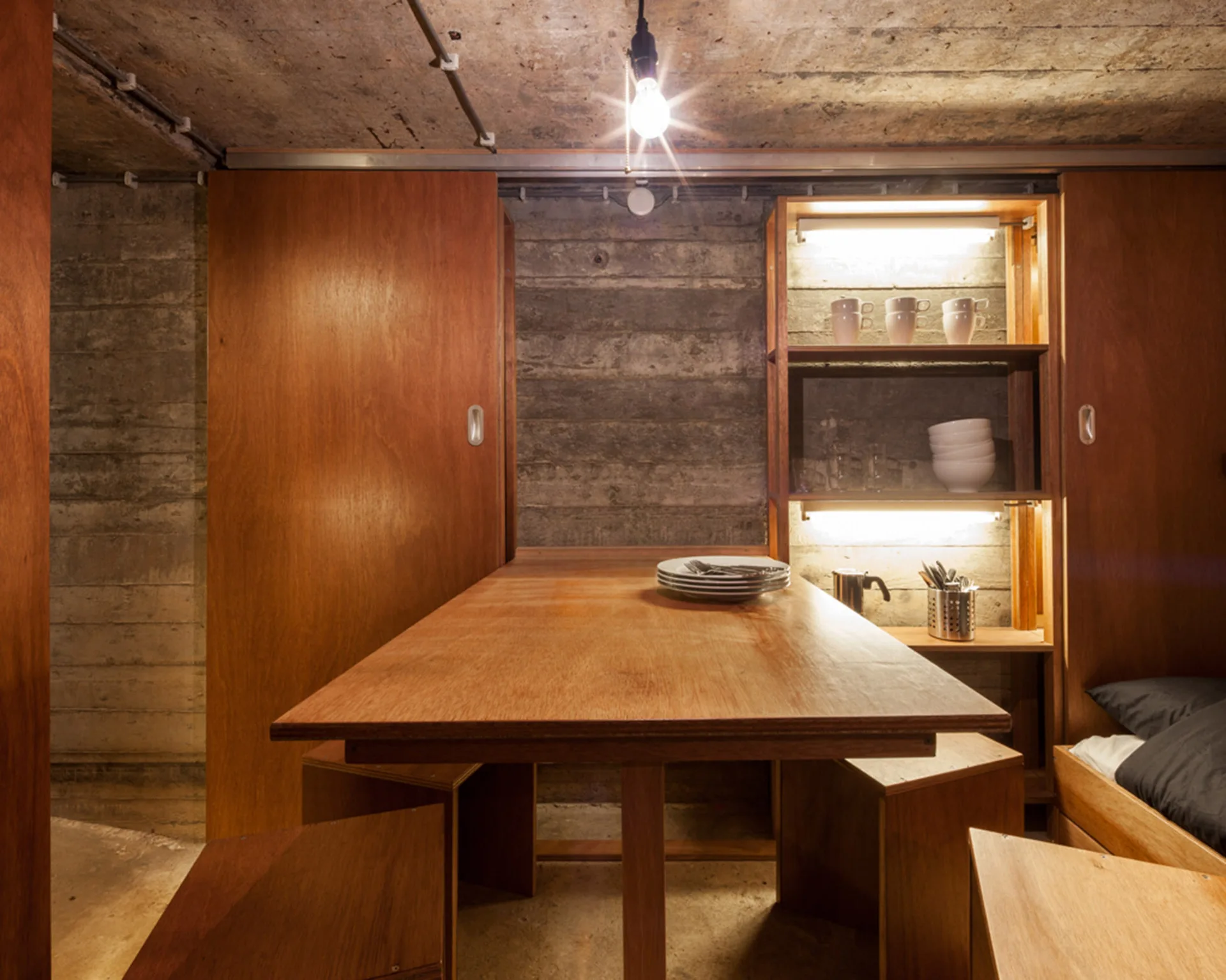 A wooden dining table and block stools set against raw concrete walls and custom built-in cabinets with integrated lighting.