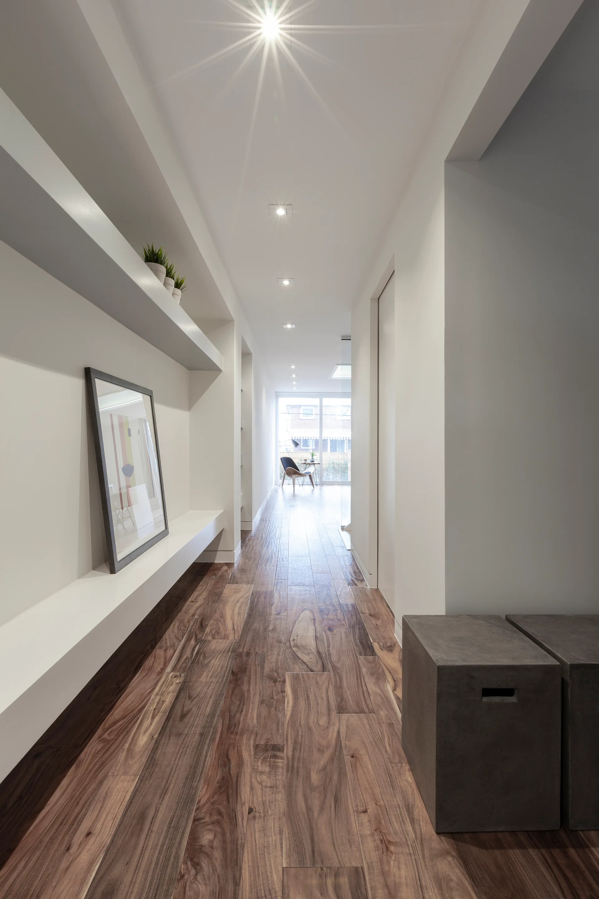 A long minimalist hallway features white walls, dark wood-grained flooring, a long built-in wall niche shelf with framed art, and recessed ceiling lights.