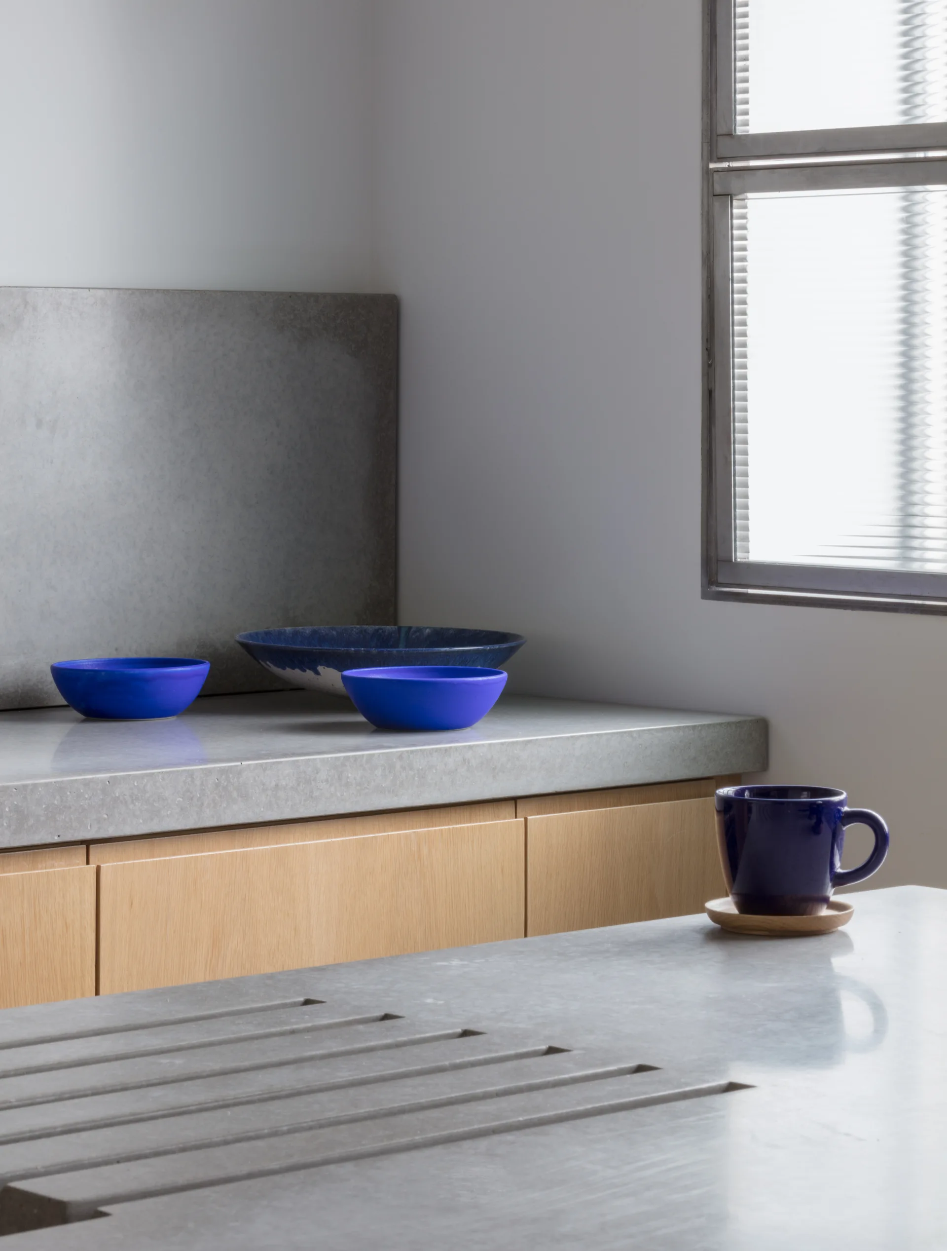 A concrete kitchen countertop features vibrant blue ceramic bowls and an integrated sink above flat light wood cabinets.