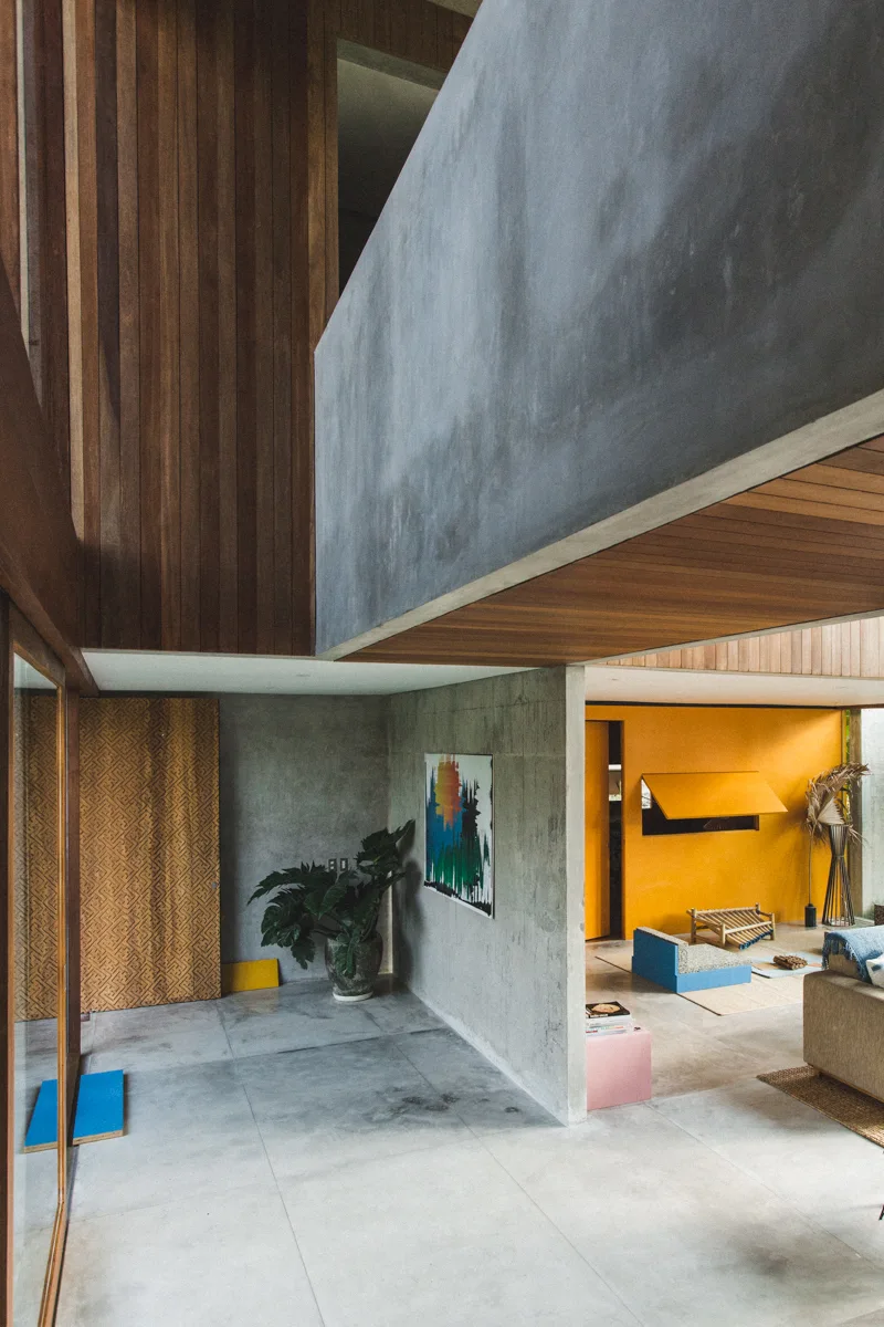 A modernist interior featuring raw concrete walls, warm wood ceiling panels, a patterned wood door, and a bright yellow accent wall.