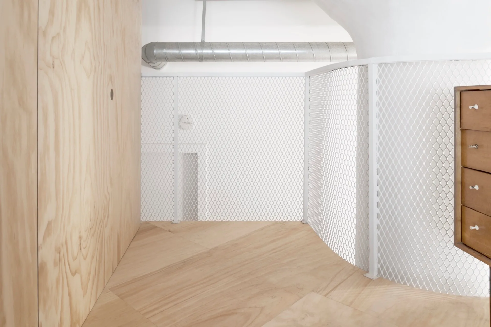 An interior loft landing featuring light wood plywood wall panels, a white metal mesh safety railing, and diagonal wood flooring.