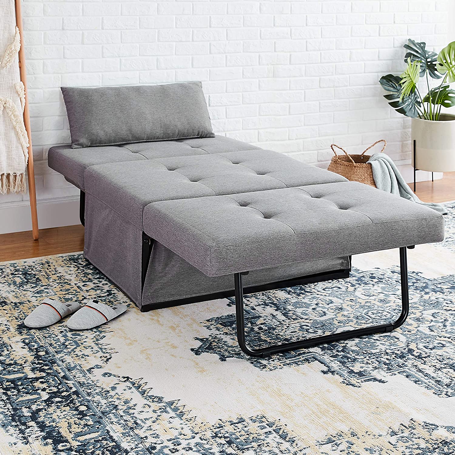 A grey tufted convertible ottoman is unfolded into a cot-sized bed on a distressed blue and cream rug against a white brick wall.