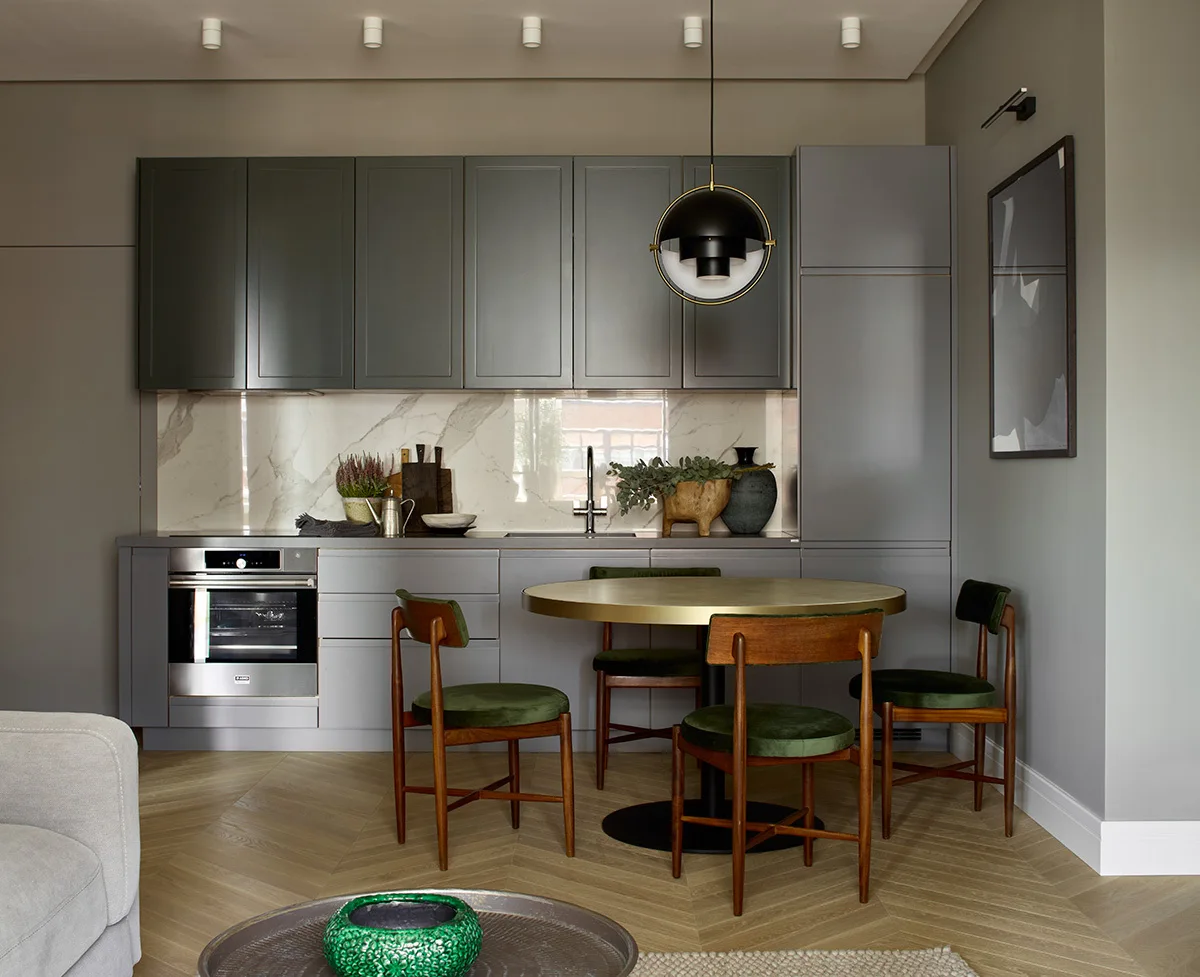 A grey kitchen features flat-panel cabinets, a white marble backsplash, a round gold dining table with green velvet chairs, and herringbone wood floors.